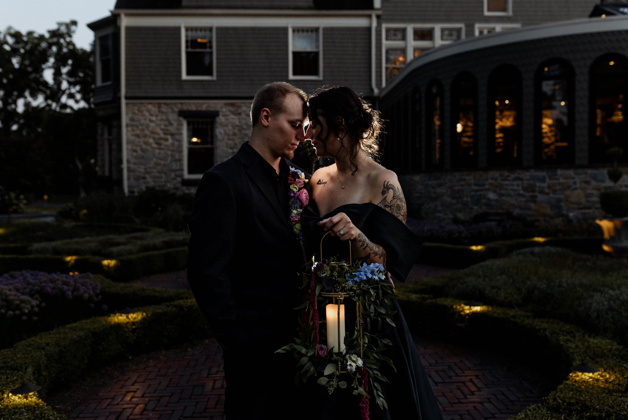 A couple stands close together in an outdoor setting at dusk, with a historic building in the background. The woman holds a lantern decorated with flowers, and they share an intimate moment with their foreheads touching.