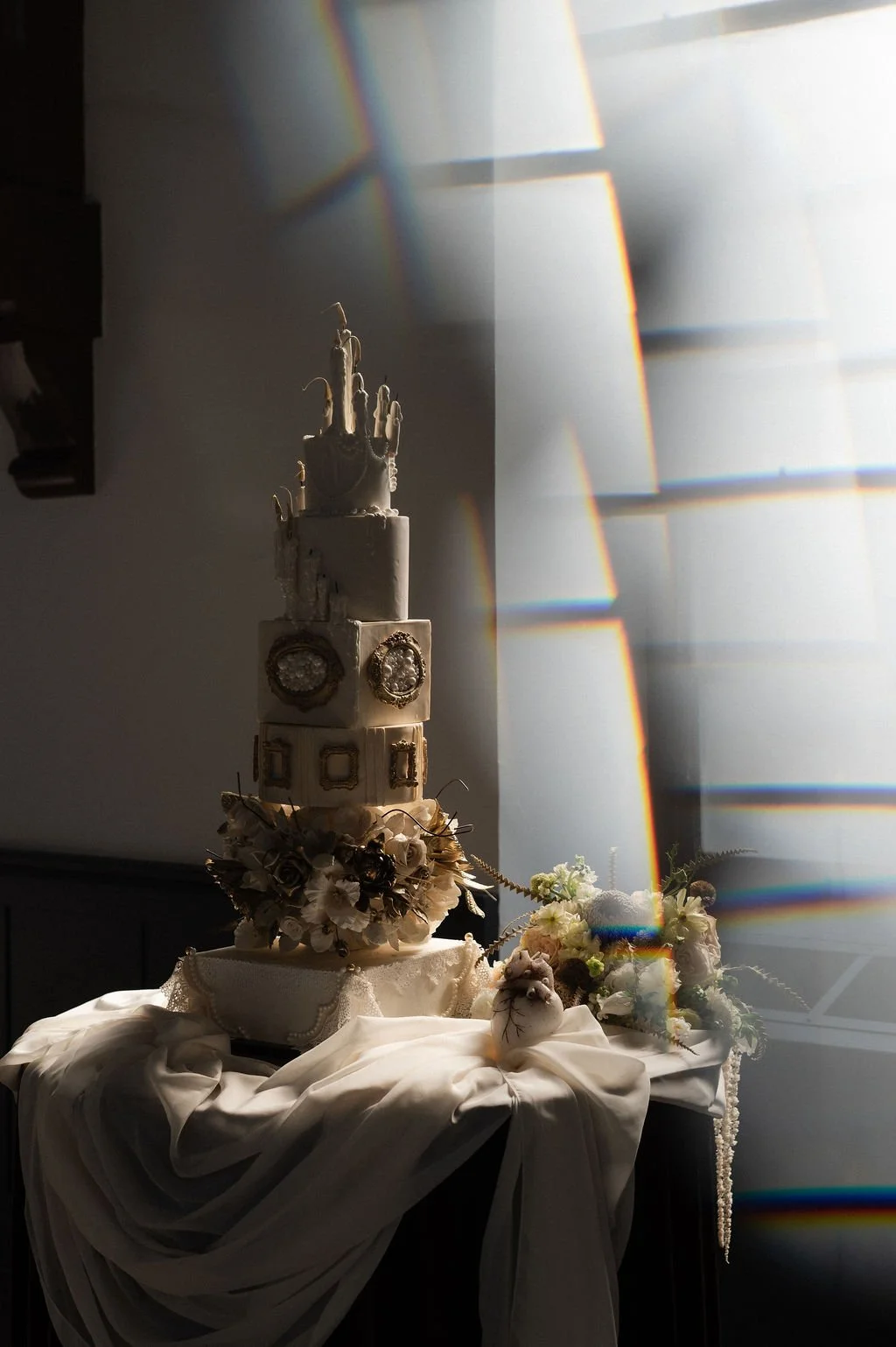 A white, multi-tiered wedding cake decorated with flowers, ribbons, and framed photographs, topped with wedding figurines. The cake is displayed on a table draped with white fabric, with a bouquet of flowers and decorative elements nearby. Light crea