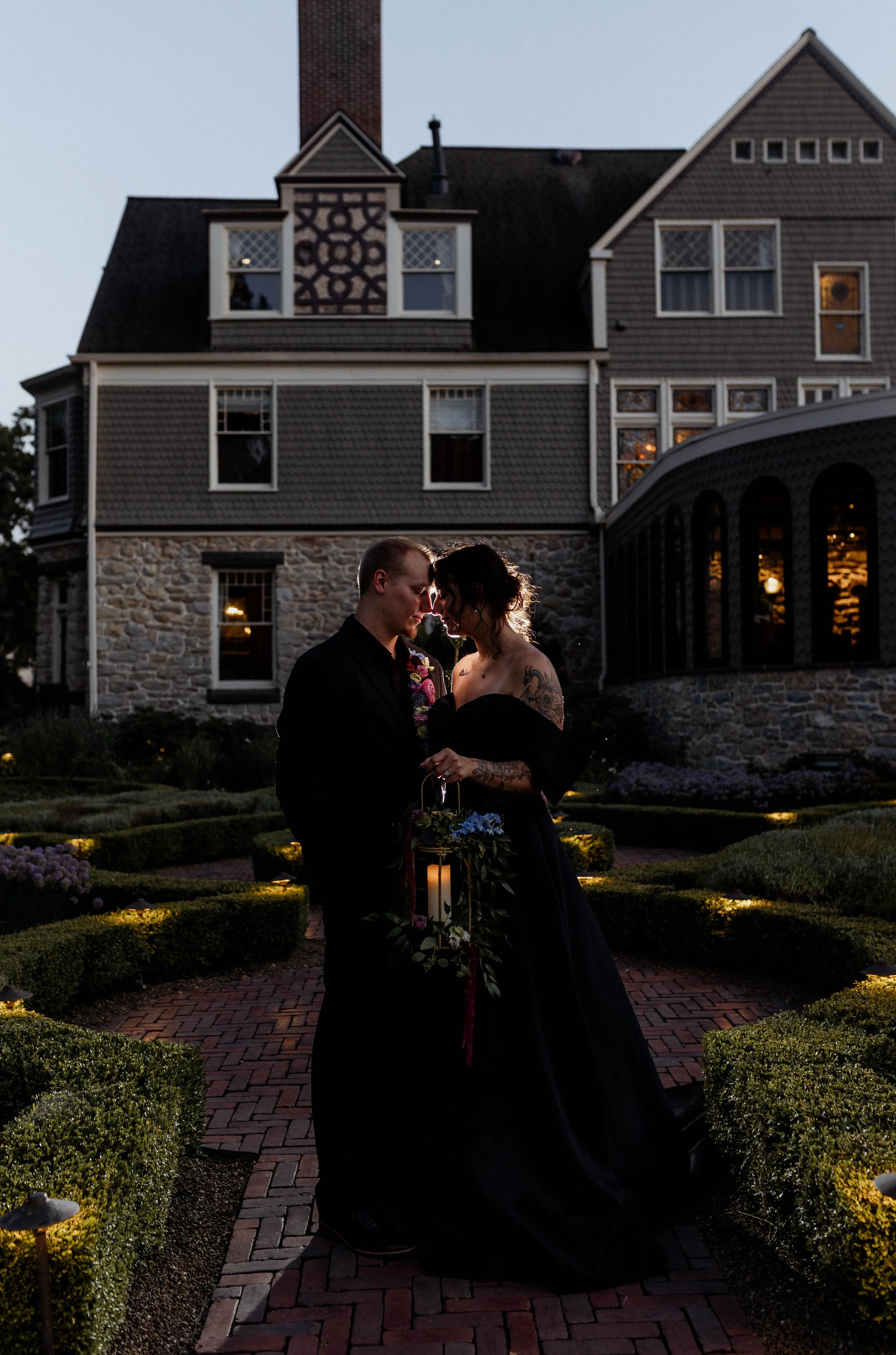 A couple dressed in black romantic clothes stands close together outside at dusk, holding a lantern and floral arrangement in front of a large, historic house with a stone and shingle exterior, lit windows, and a tall brick chimney.
