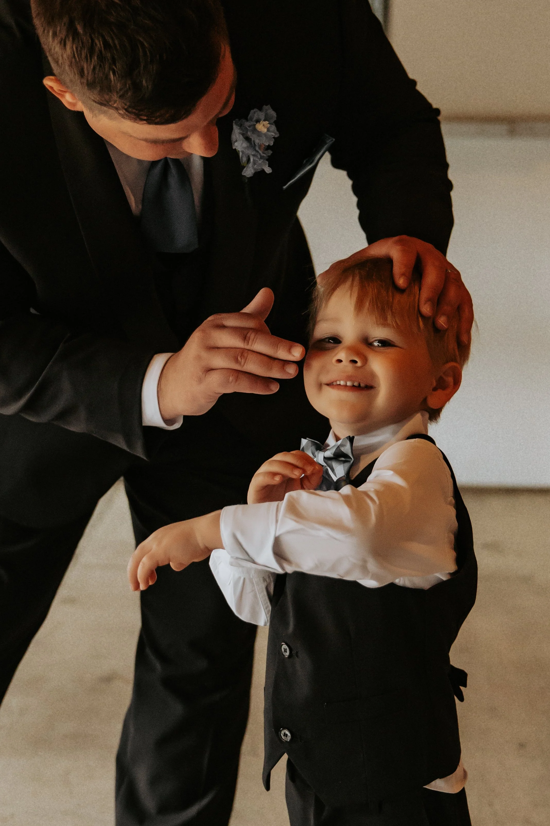 A man in a black suit gently touches a young boy's face, who is smiling and dressed in a white shirt and black vest with a bow tie, during a formal event.