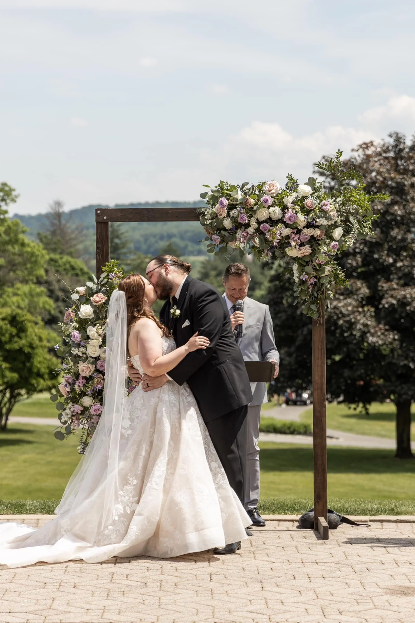A bride and groom share a kiss during their outdoor wedding ceremony, with an officiant standing behind them holding a microphone and a black folder. They are under a wooden arch decorated with a large floral arrangement, on a sunny day in a park or 