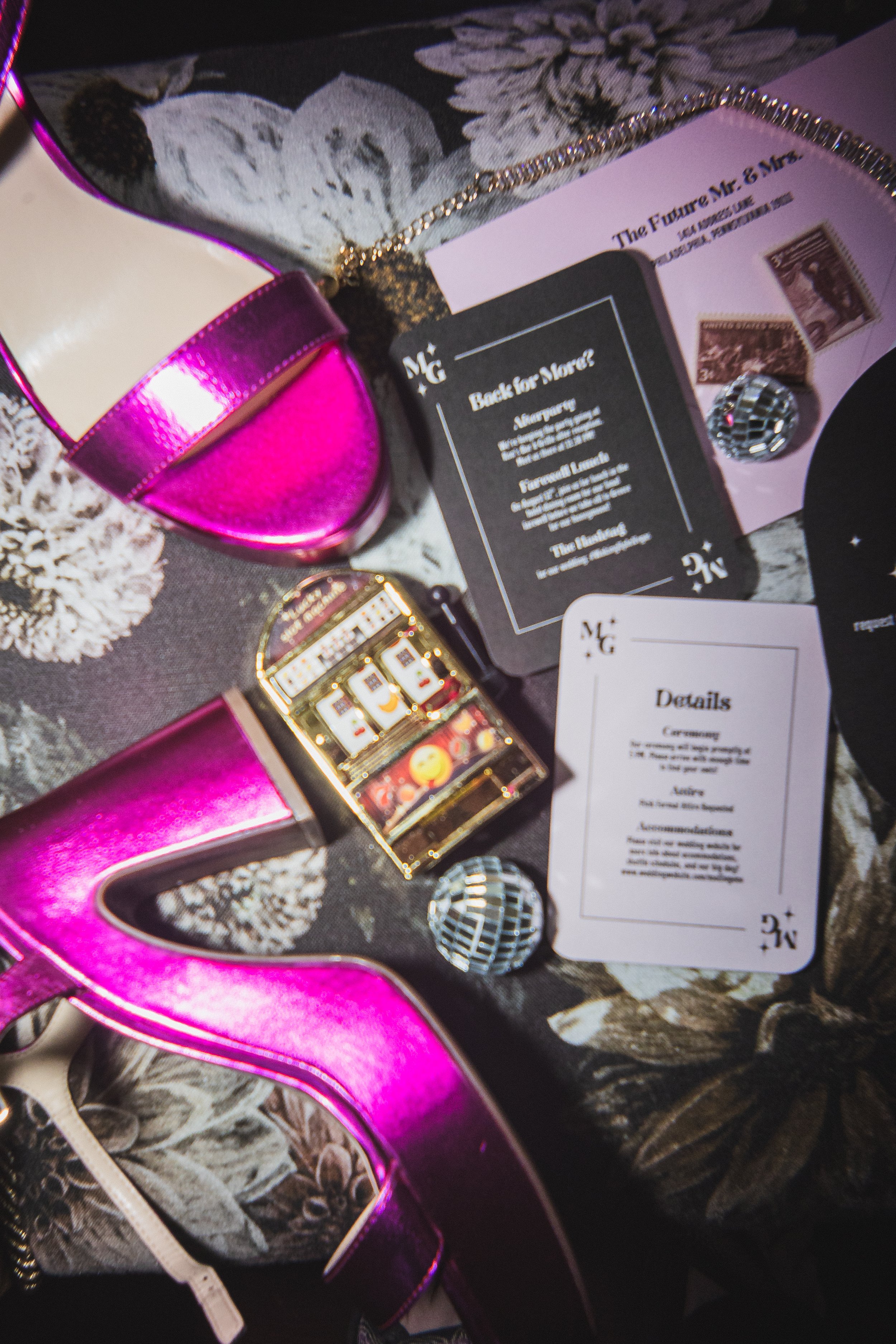 Pink high-heeled shoes, casino chips, playing cards, and a miniature slot machine on a floral-patterned tablecloth.