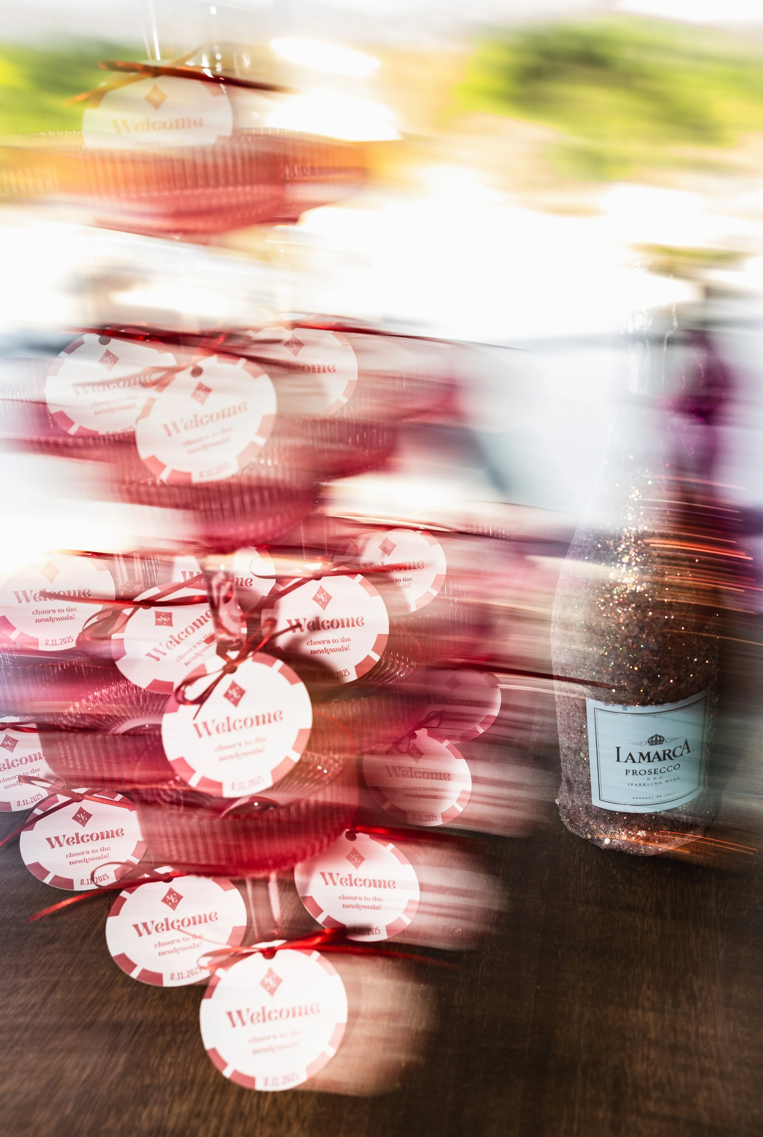 Stacked red gift boxes with welcome tags and a bottle of Lamarca Prosecco with glitter on the right.