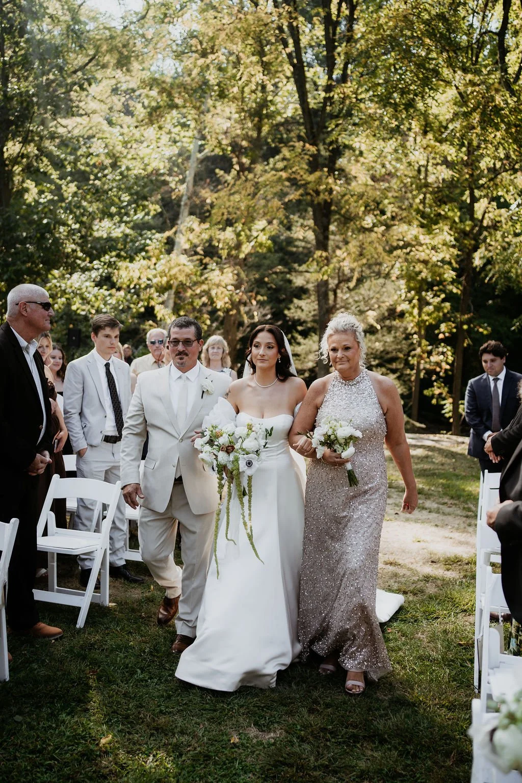 A bride in a white wedding gown walking down the outdoor aisle with her parents, surrounded by wedding guests on a sunny day in a wooded area.