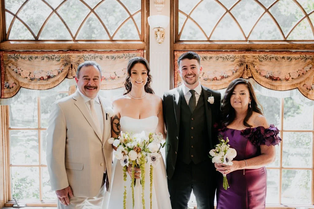 Wedding celebration indoors with four people, two men and two women, standing in front of large windows with floral curtains, smiling for the camera.