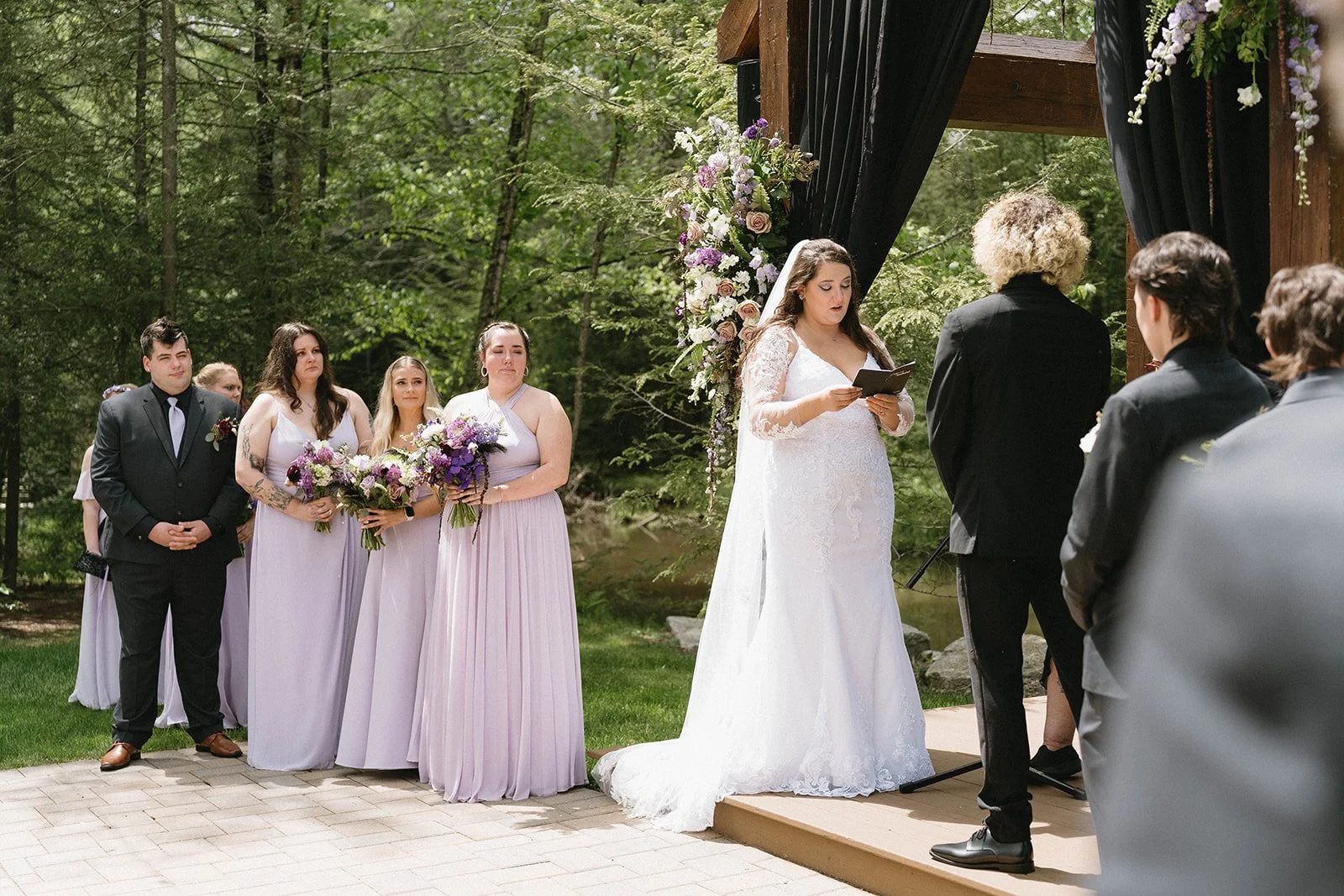 A bride in a white wedding gown reading vows during an outdoor wedding ceremony, with bridesmaids in lavender dresses holding bouquets and groomsmen in black tuxedos, in a wooded setting with floral decorations.