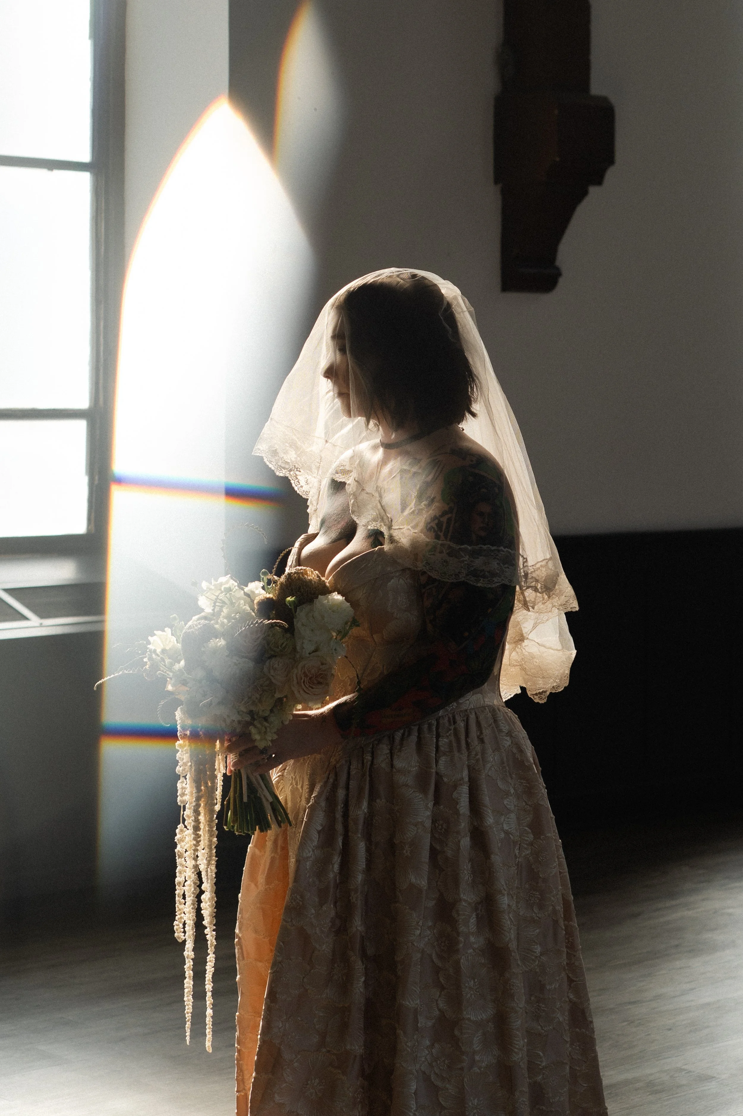 A woman in a wedding dress and veil holding a bouquet of flowers, standing in front of a window with sunlight and a rainbow lens flare.