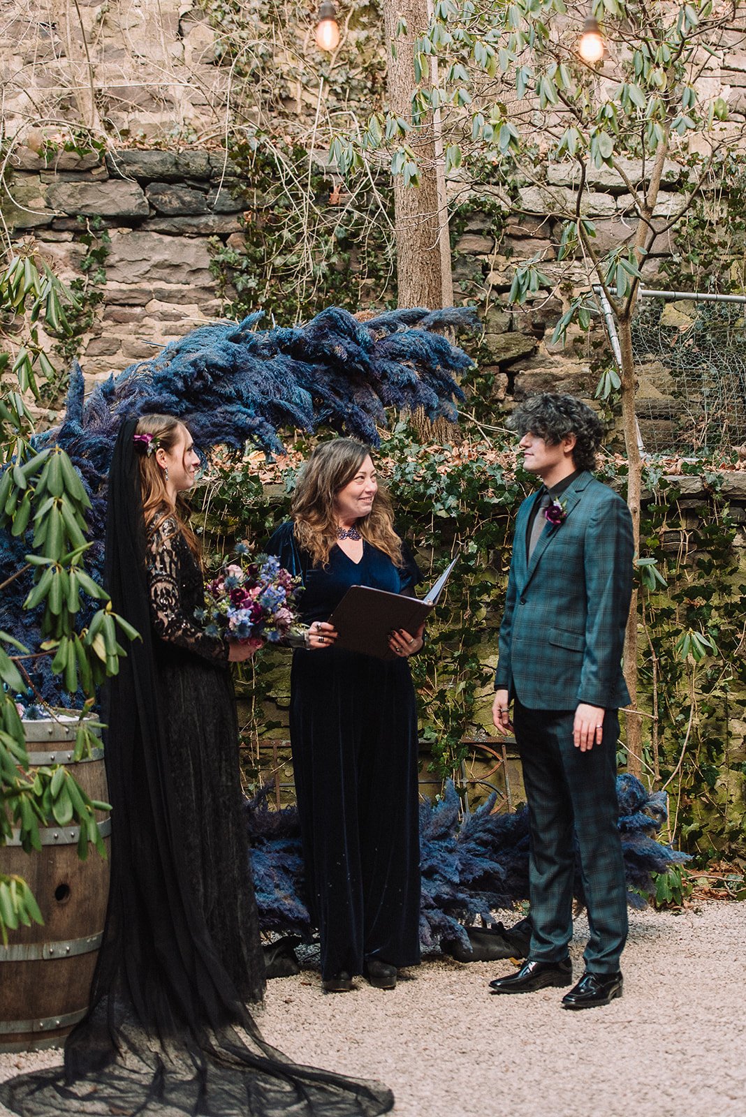 A wedding ceremony with two women and a man outdoors, surrounded by green foliage and string lights. One woman holds flowers; the other is reading vows to a man in a checked suit. There is a large blue floral arrangement in the background.