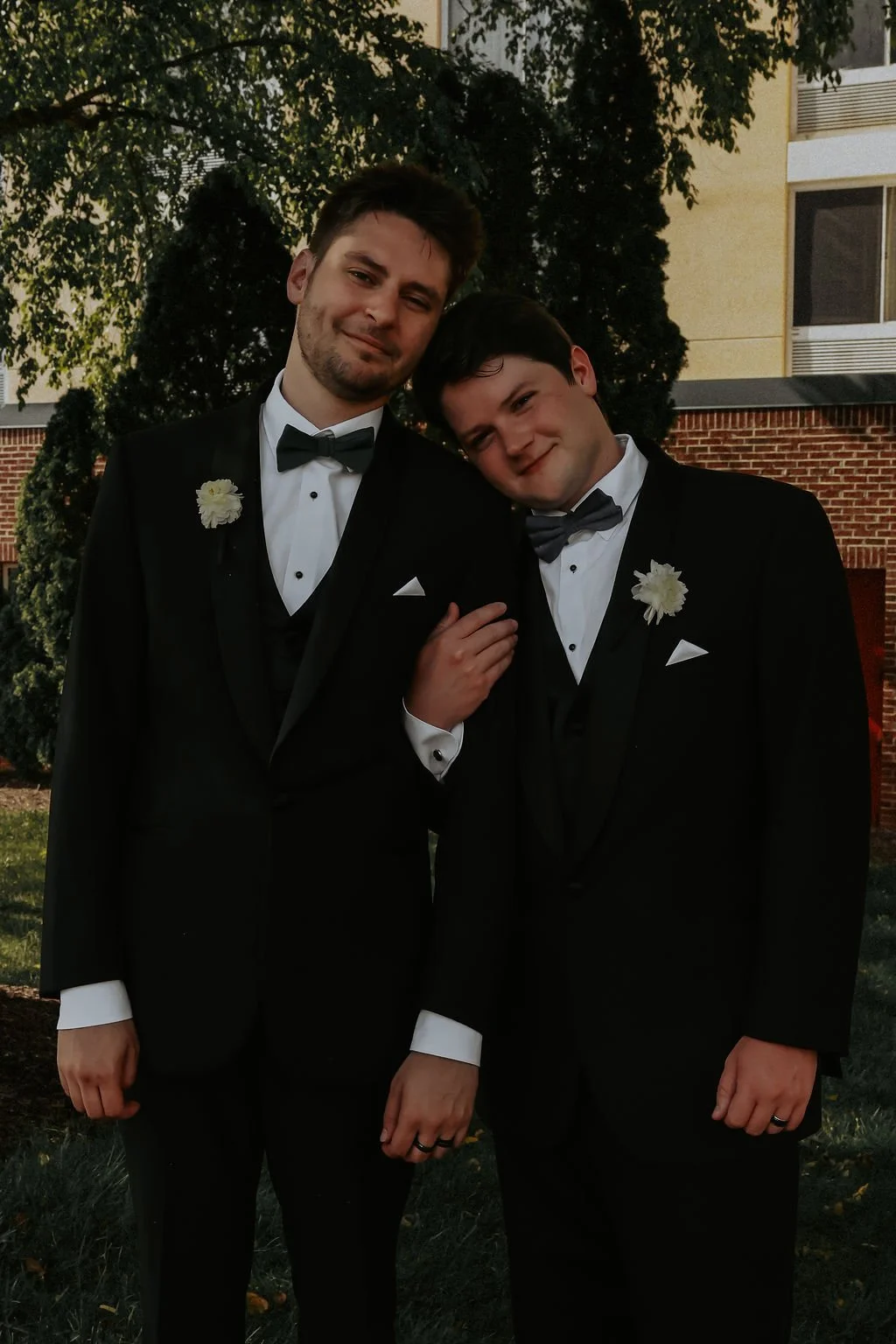 Two young men in tuxedos with boutonnières, standing close together outdoors during daytime, smiling at the camera.