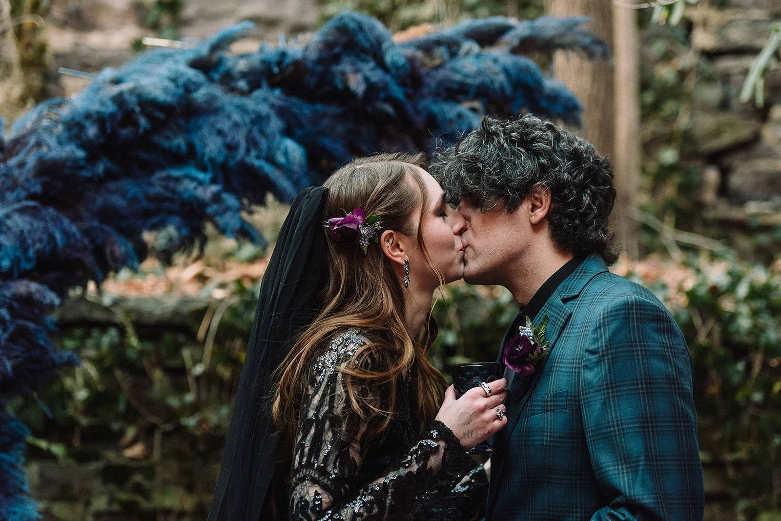 A woman and a man kissing at an outdoor wedding, with the woman holding a small black cup, surrounded by a wooden and natural setting.
