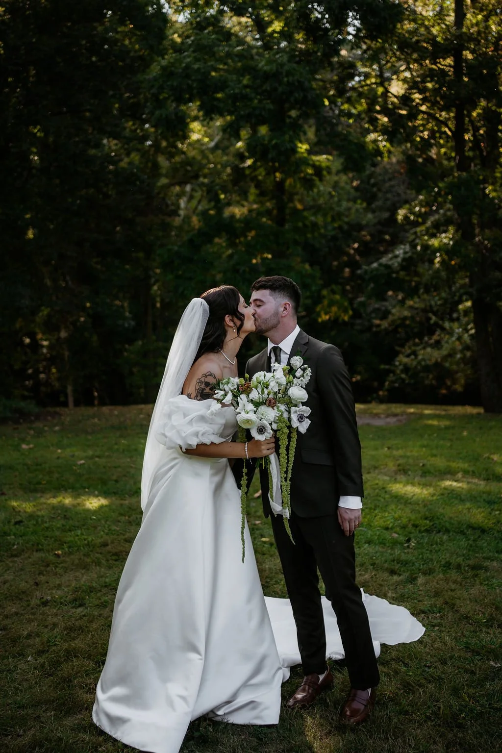 A bride and groom share a kiss outdoors in a park with trees in the background, the bride wearing a white wedding gown with puffed sleeves and a veil, and the groom in a black suit with a white shirt and black tie, holding a bouquet of white and gree