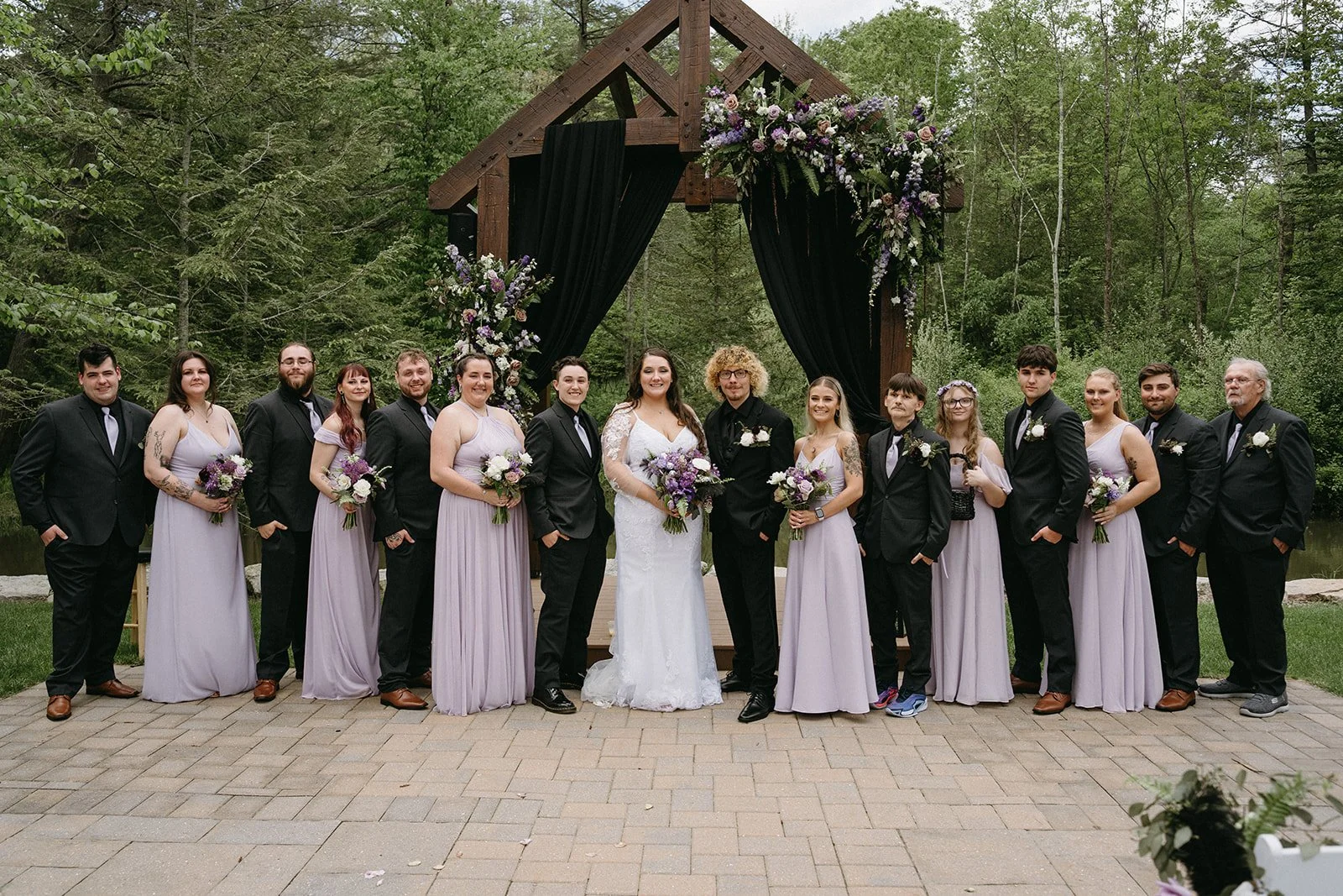A wedding party standing outdoors in front of a wooden arch decorated with flowers and black curtains. The group includes the bride in a white gown, the groom, bridesmaids in lavender dresses, and groomsmen in black suits, all holding bouquets or wit
