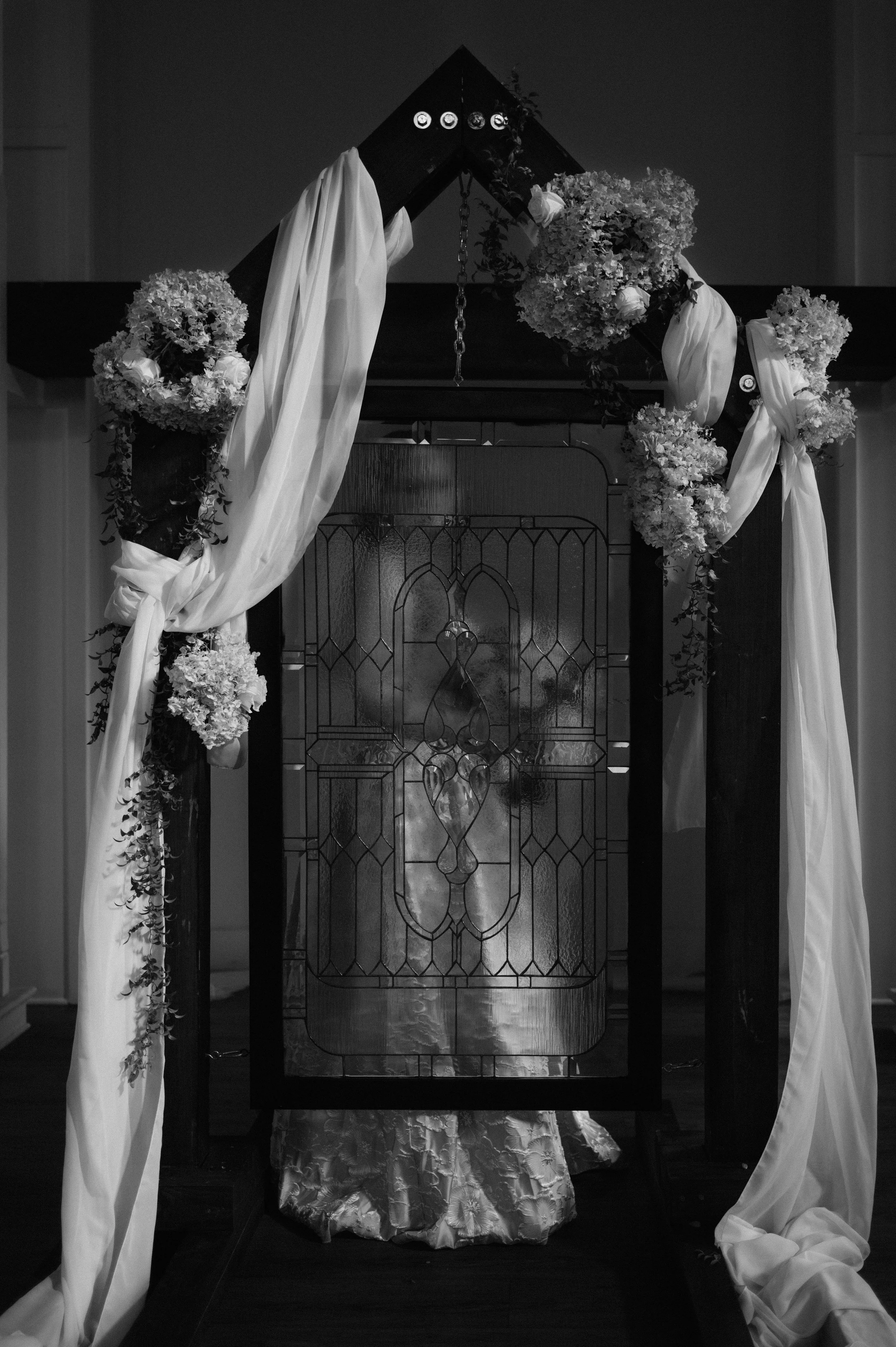 A wedding altar decorated with flowers, white fabric, and a stained glass window in the center.