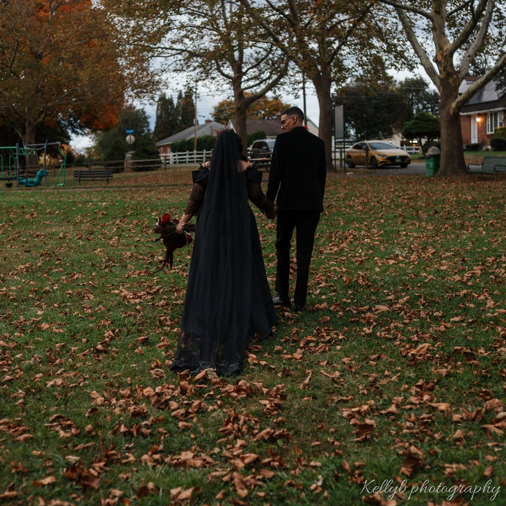A couple dressed in formal attire walking hand in hand through a park with fallen autumn leaves, trees, and houses in the background.
