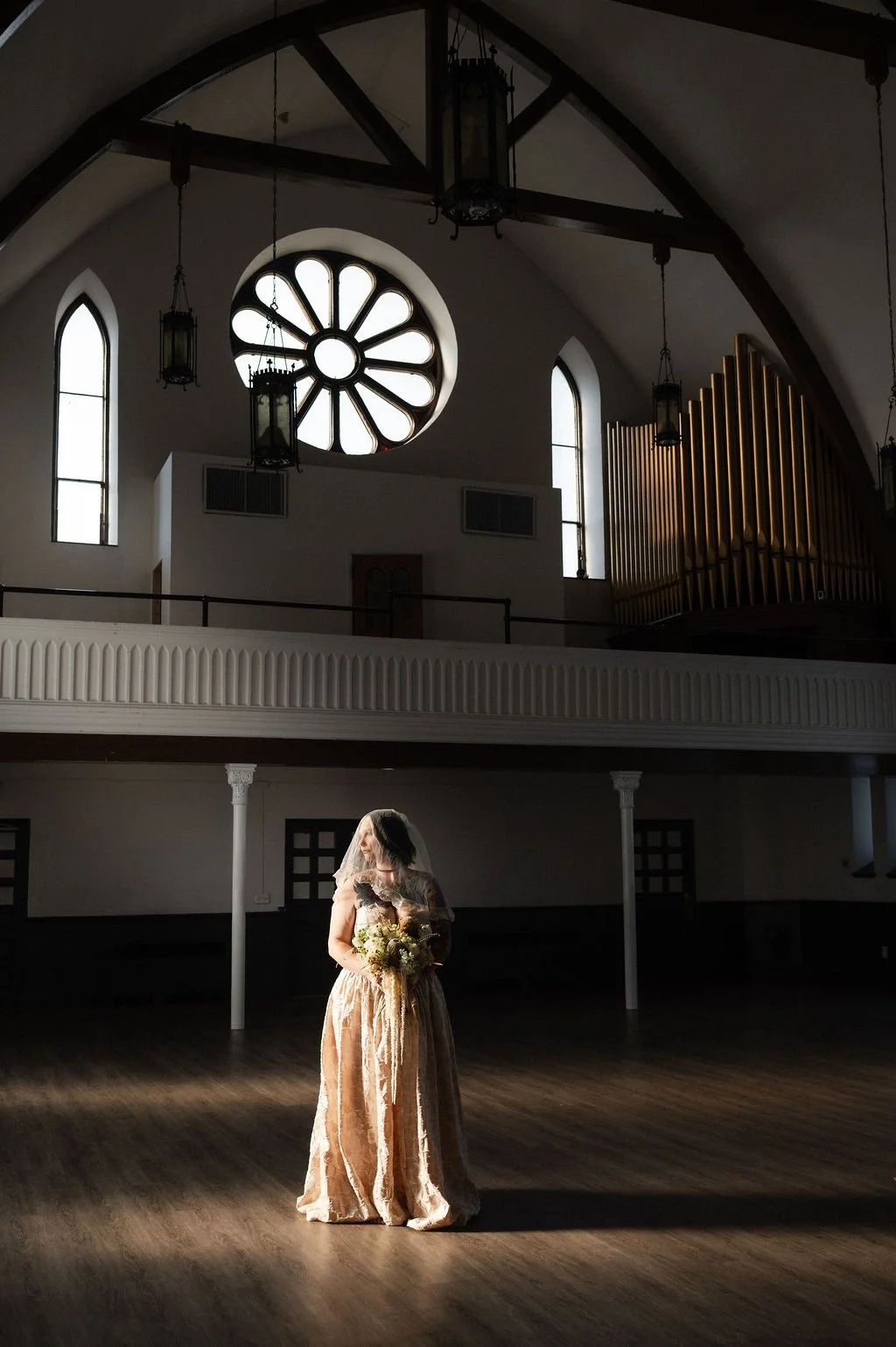 A bride holding a bouquet in a large, dimly lit church with tall arched windows and a large round stained glass window above her.