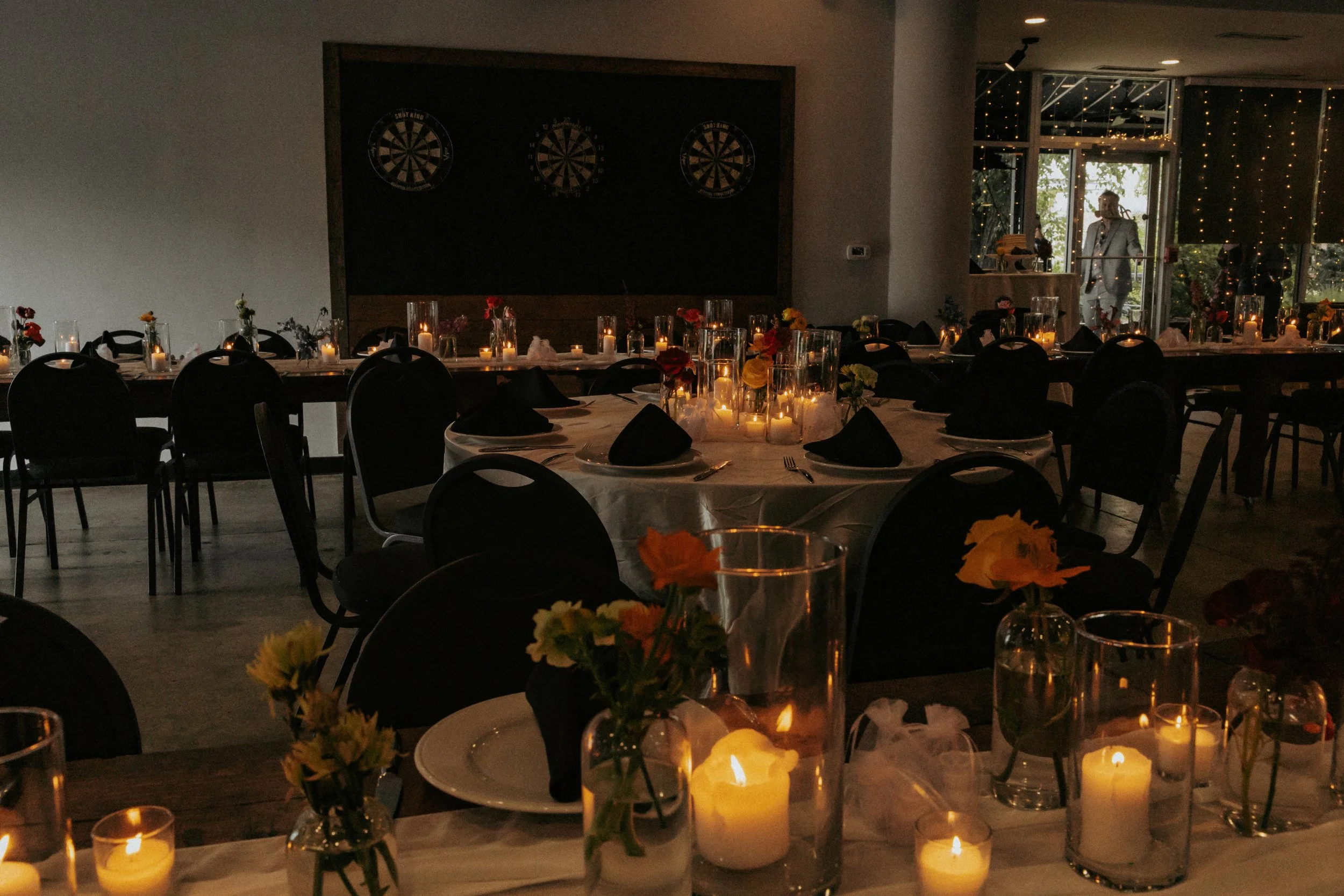 Indoor event space with tables decorated with candles and flowers, prepared for a formal dinner or celebration, with dartboards on the wall and a person standing near the entrance.