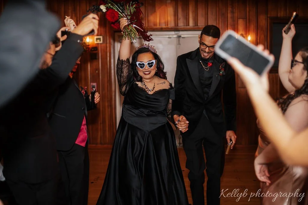 A bride and groom holding hands and smiling, surrounded by wedding guests taking photos, at a wedding reception with wood-paneled walls and floral decorations.