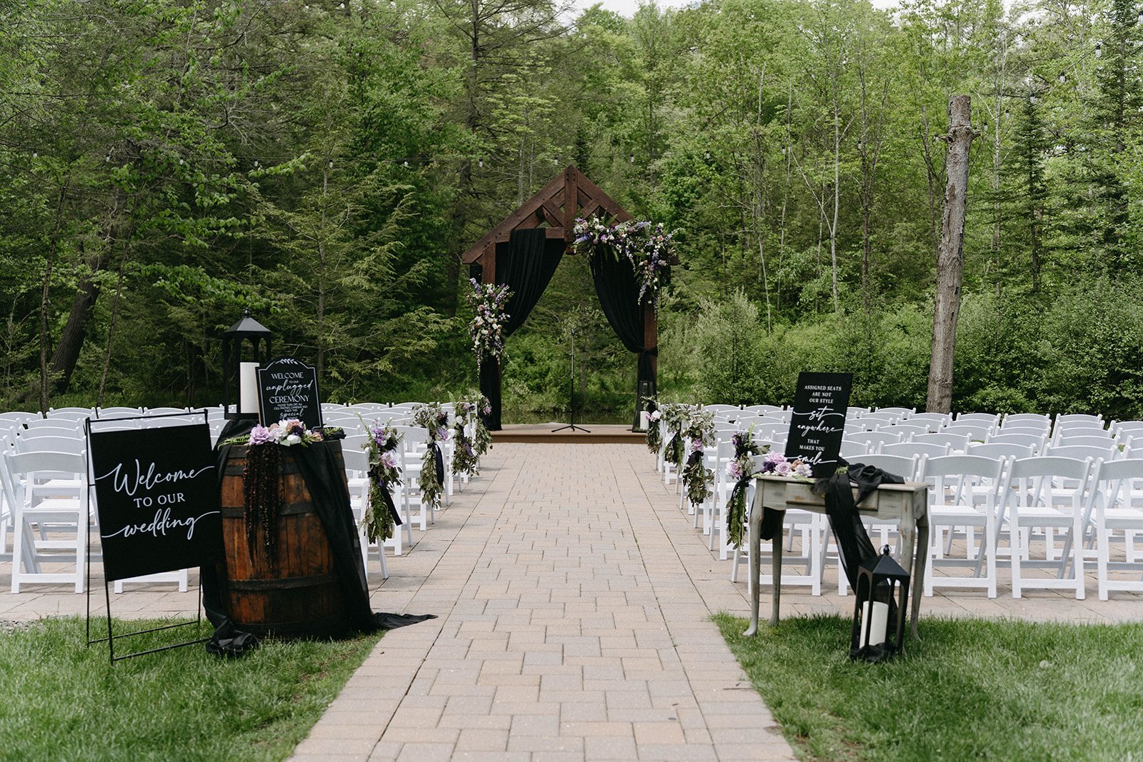 An outdoor wedding setup with white chairs arranged facing a wooden arch decorated with flowers, on a paved aisle in a forested area.