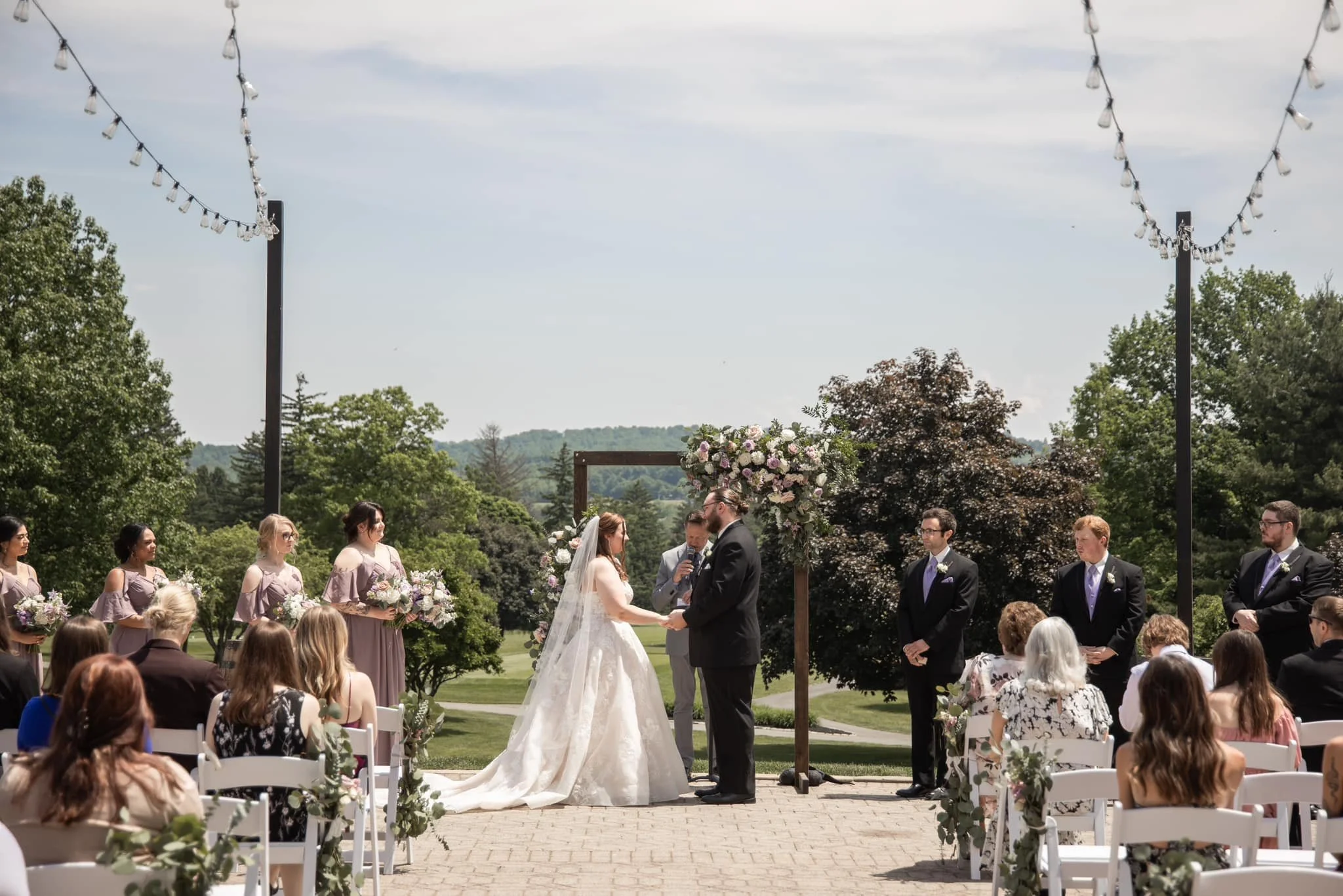 Outdoor wedding ceremony with the bride and groom holding hands, officiant in the background, bridesmaids and groomsmen standing on either side, guests seated, decorated with floral arrangements and string lights, greenery and trees in the background