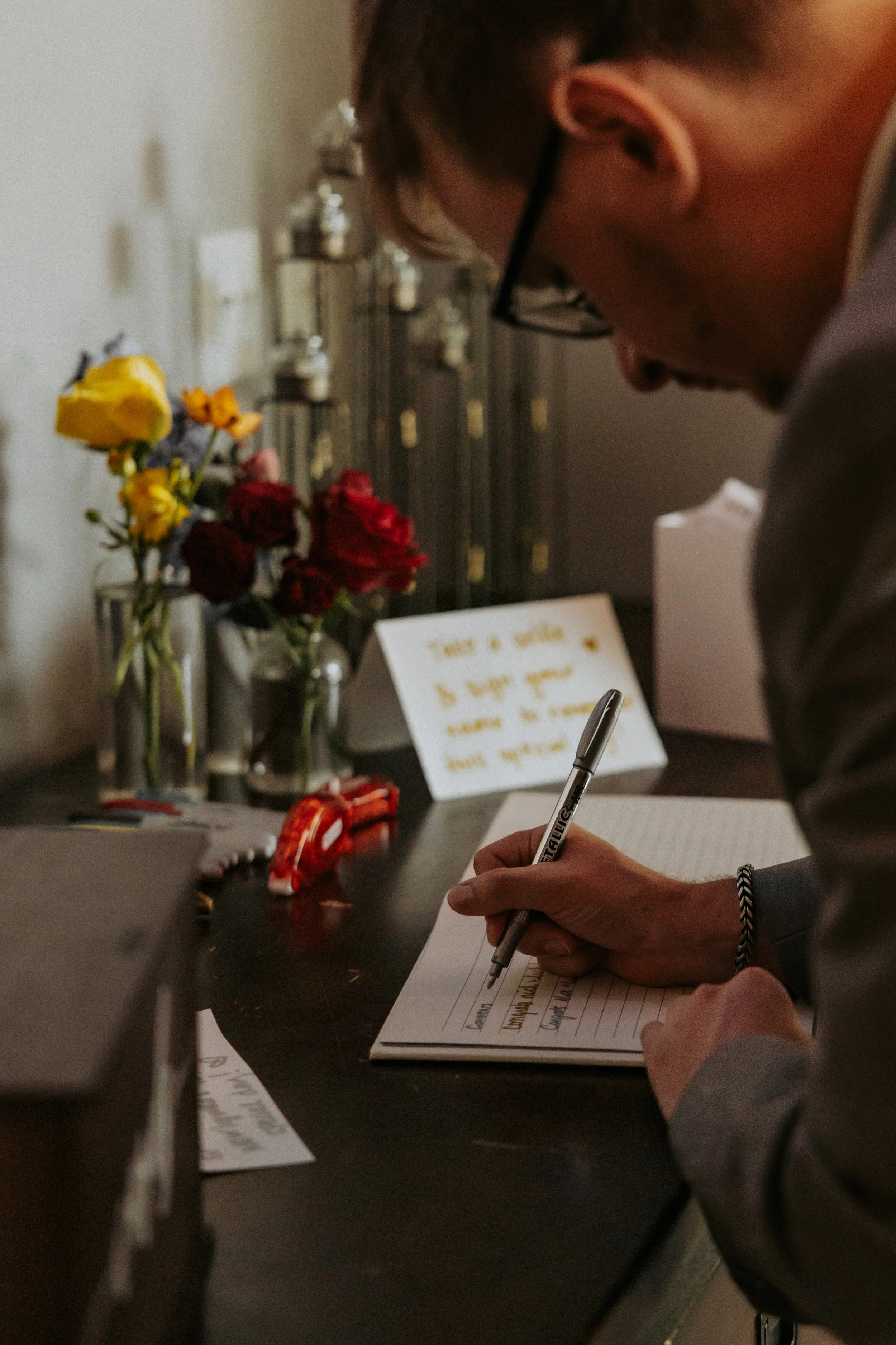 A person writing on a notepad at a desk with flowers, notes, and office supplies.