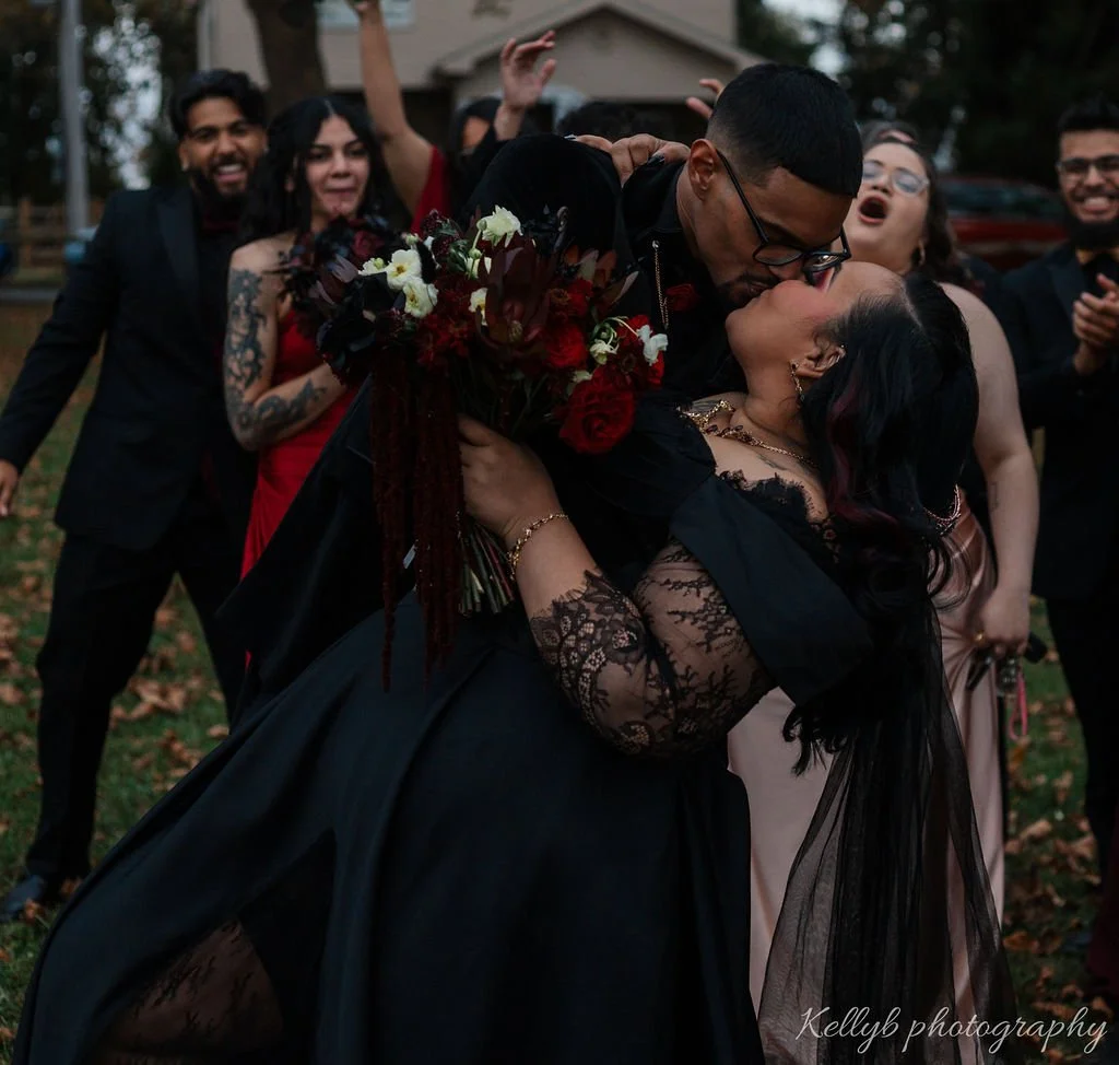 A couple sharing a kiss during a celebration outdoors, surrounded by people clapping and smiling.