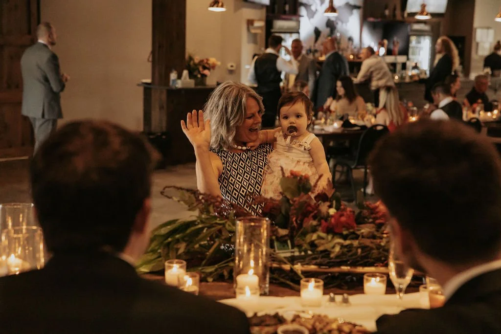Woman holding a young girl at a social gathering, with candles and floral arrangements on the table, other guests mingling in the background.