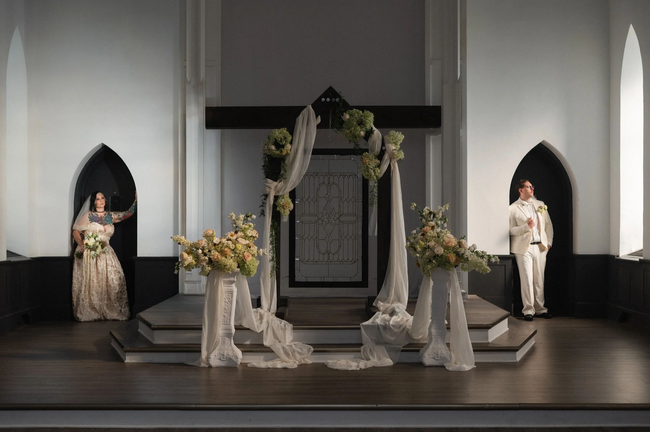 Empty wedding altar decorated with white drapes and floral arrangements, two individuals standing on each side inside a church with white walls and arched windows.