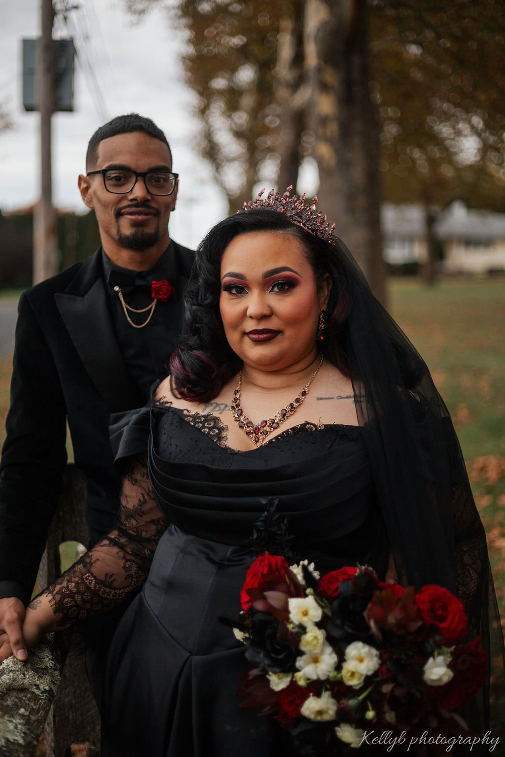 A woman wearing a black dress, a tiara, and jewelry holds a bouquet of red, white, and black flowers. A man in glasses, wearing a black suit with a red rose and gold chains, stands behind her outdoors on a fall day.
