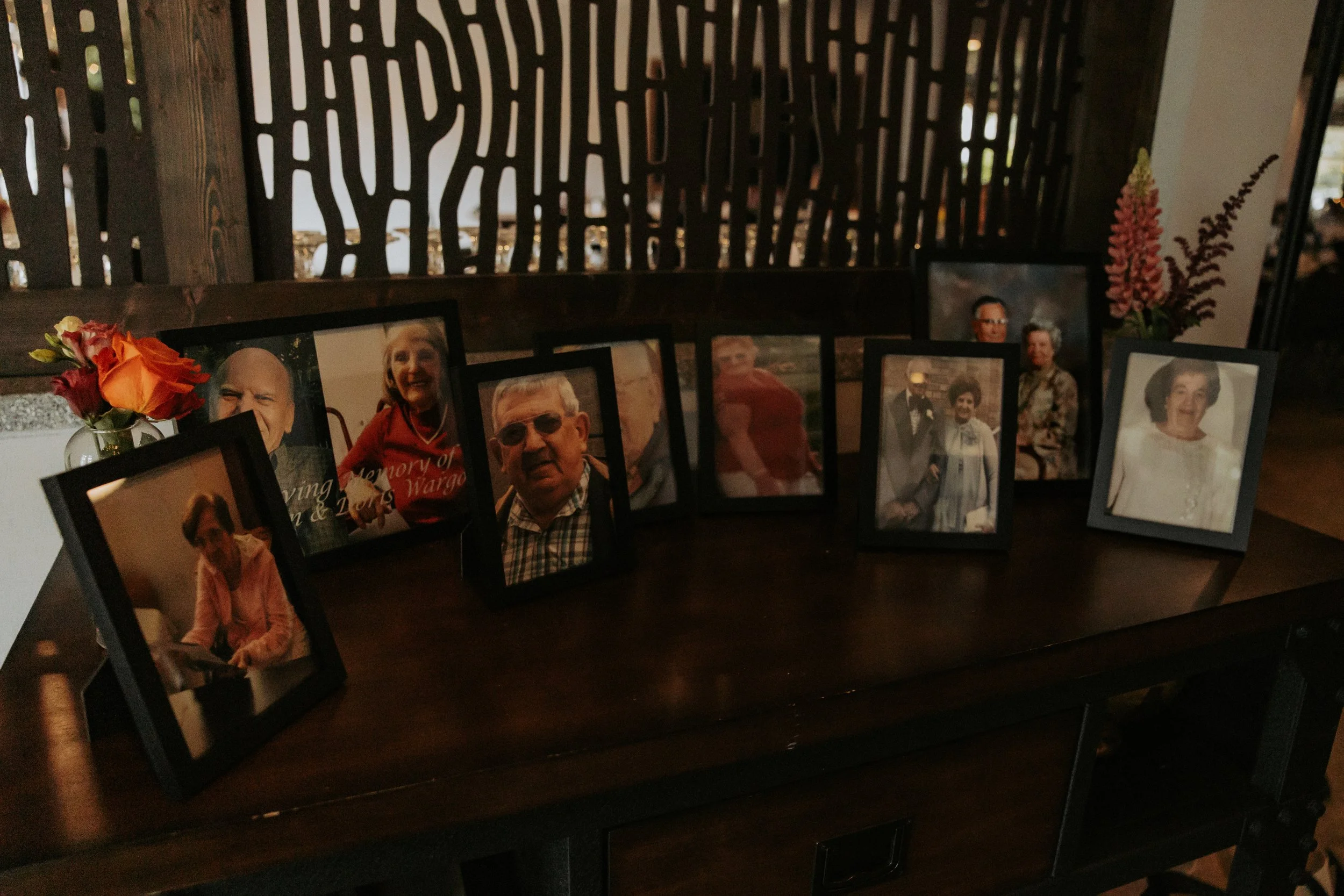 A collection of framed photographs of elderly individuals displayed on a dark wooden table with two small vases holding flowers, set against a decorative wooden partition in a warmly lit room.