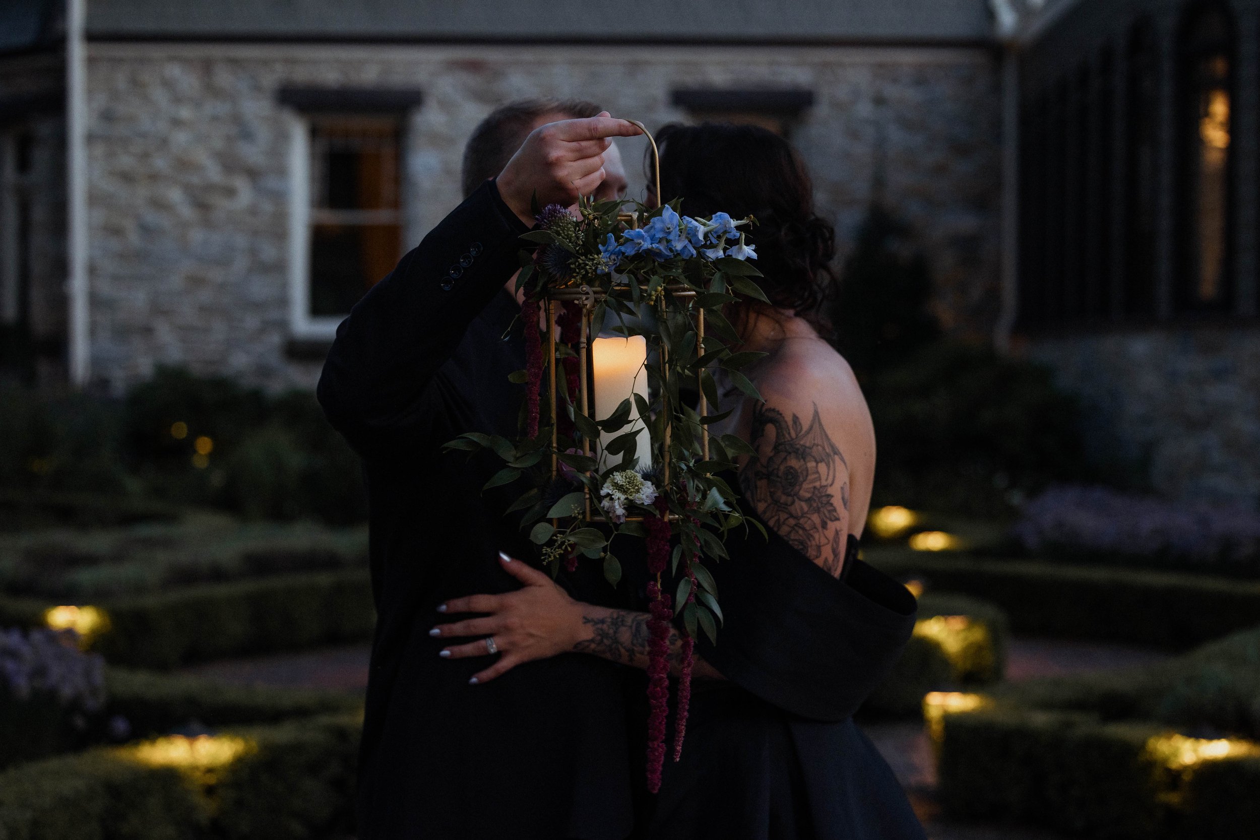 A couple dances outdoors during the evening, with the man holding a floral lantern with a lit candle, and the woman with dark hair and tattoos on her shoulder, wearing an off-shoulder black dress. The background features a stone building and garden w