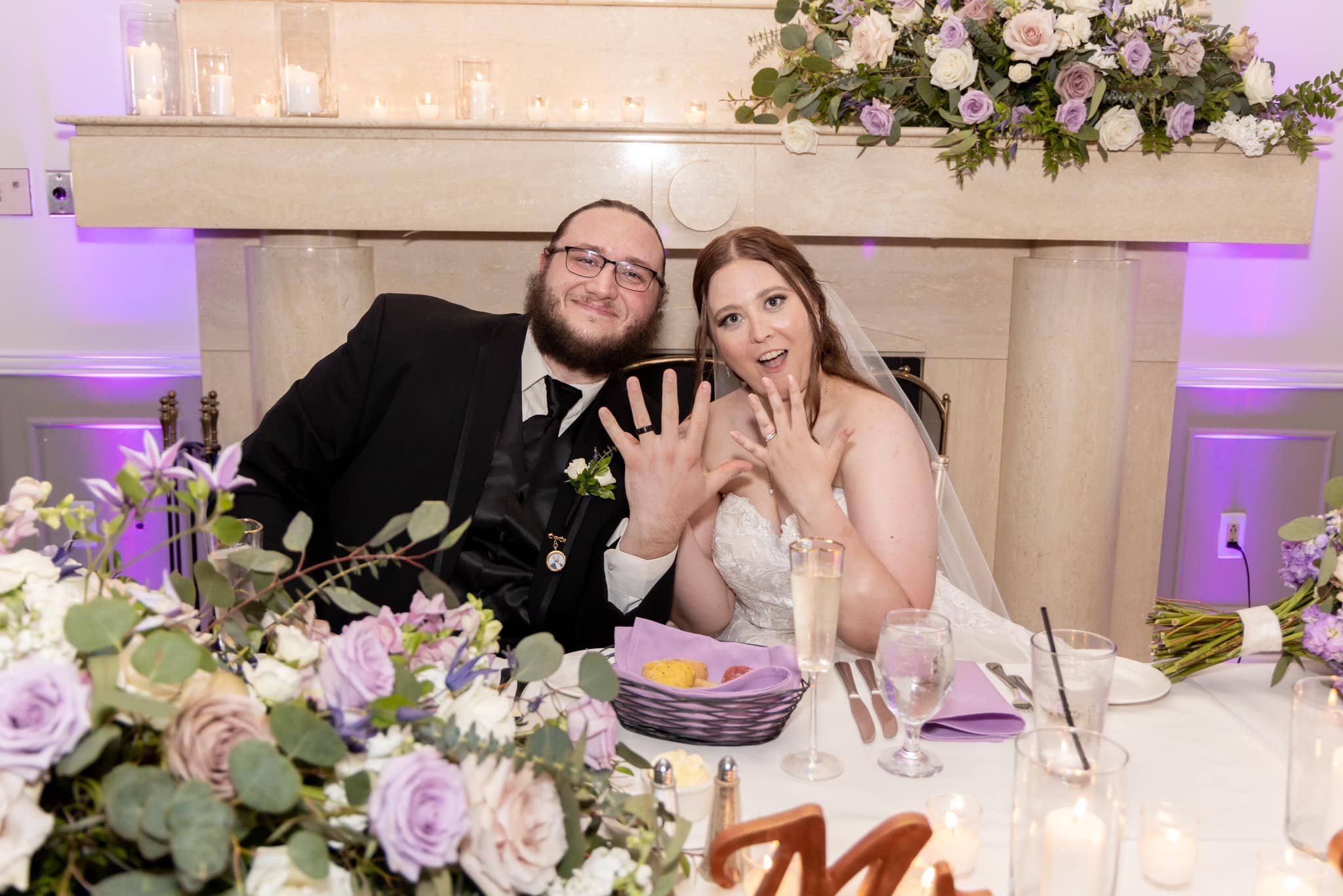 A newlywed couple at their wedding reception, displaying their wedding rings on their open hands, sitting at a decorated table with floral arrangements and candles in the background.