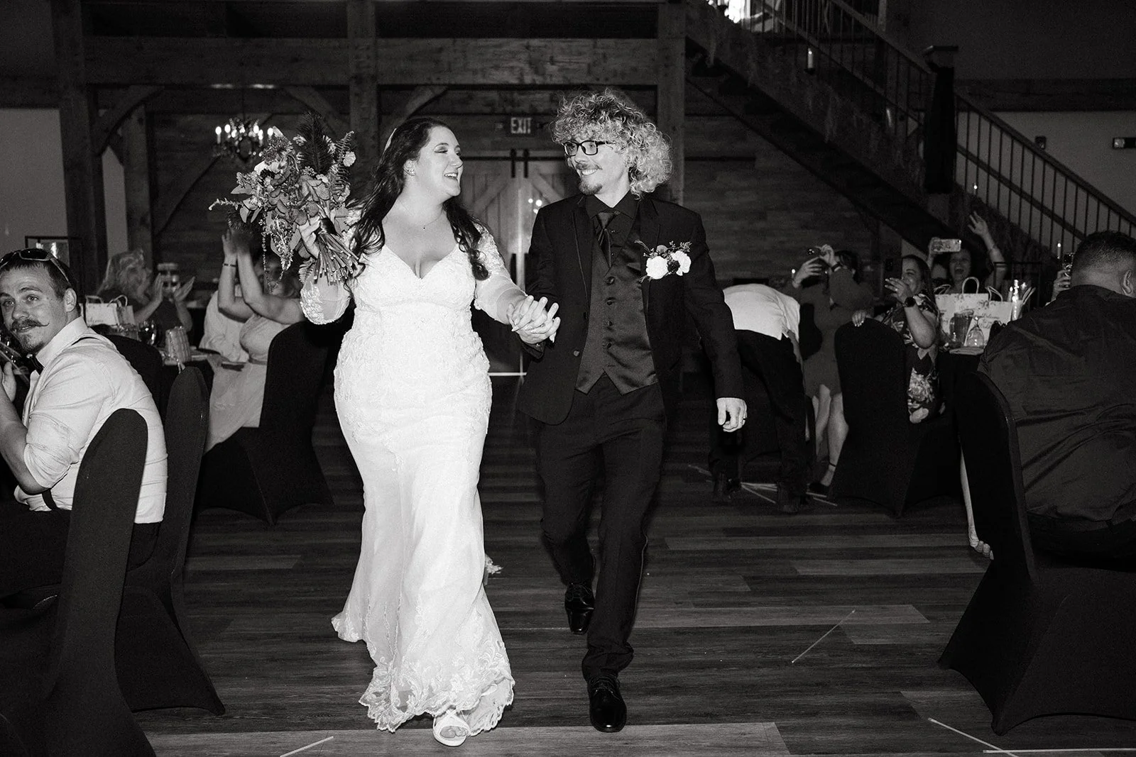 A bride and groom walking hand in hand at their wedding reception, smiling at each other. The bride is holding a large bouquet of flowers, and the groom is wearing a dark suit with a boutonniere. Guests are seated at tables around them in a rustic wo