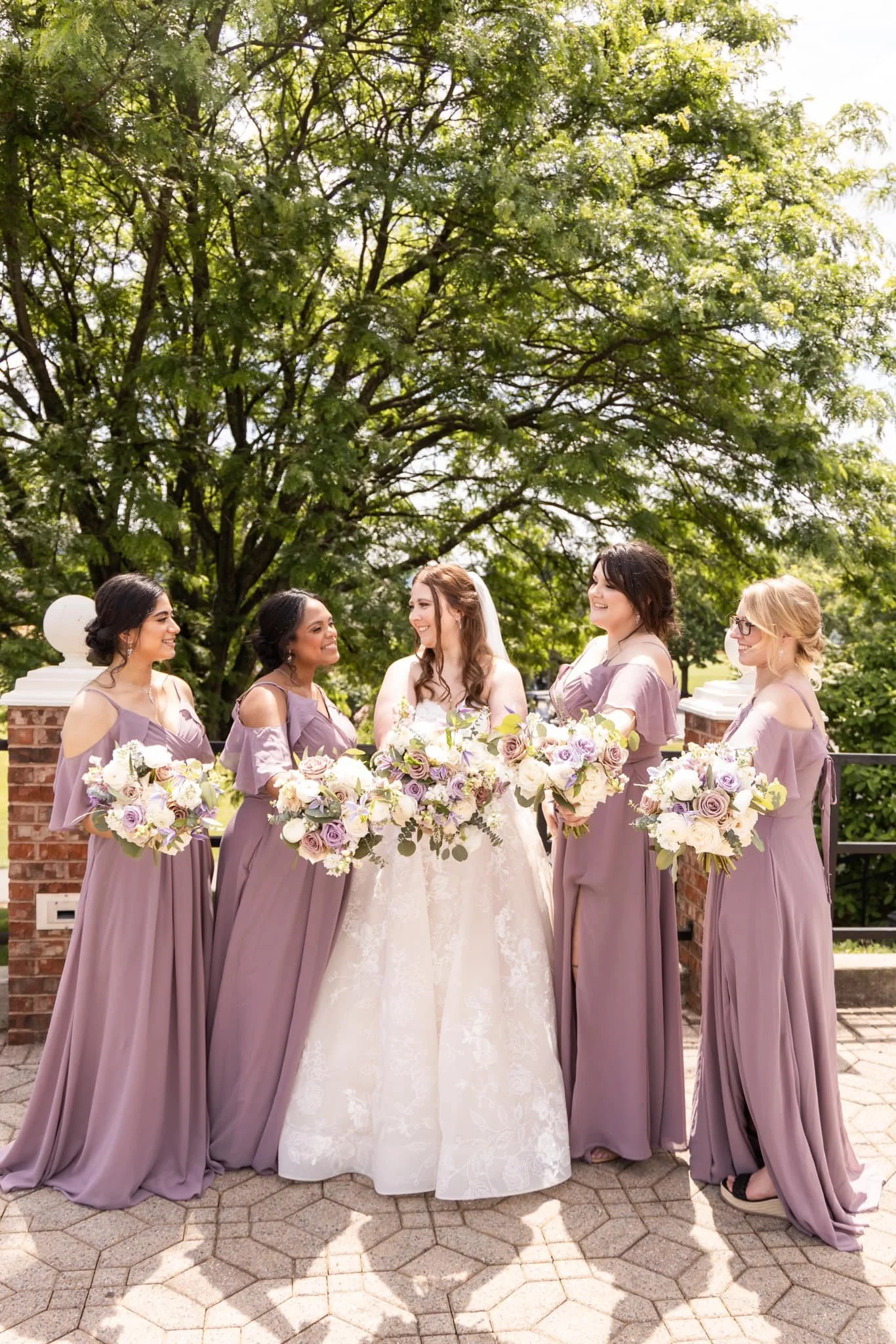 A bride in a white wedding gown with brown hair and five bridesmaids in lavender dresses holding bouquets, standing outside in front of a large green tree and brick wall.