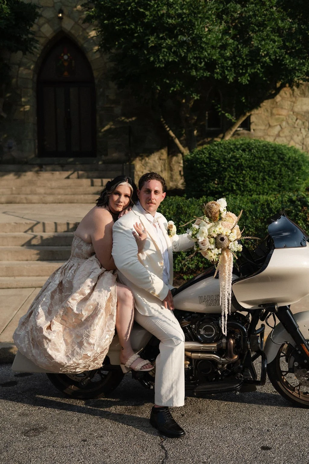 A couple in wedding attire posing on a white Harley Davidson motorcycle decorated with a bouquet of flowers, in front of a stone building with steps and greenery.