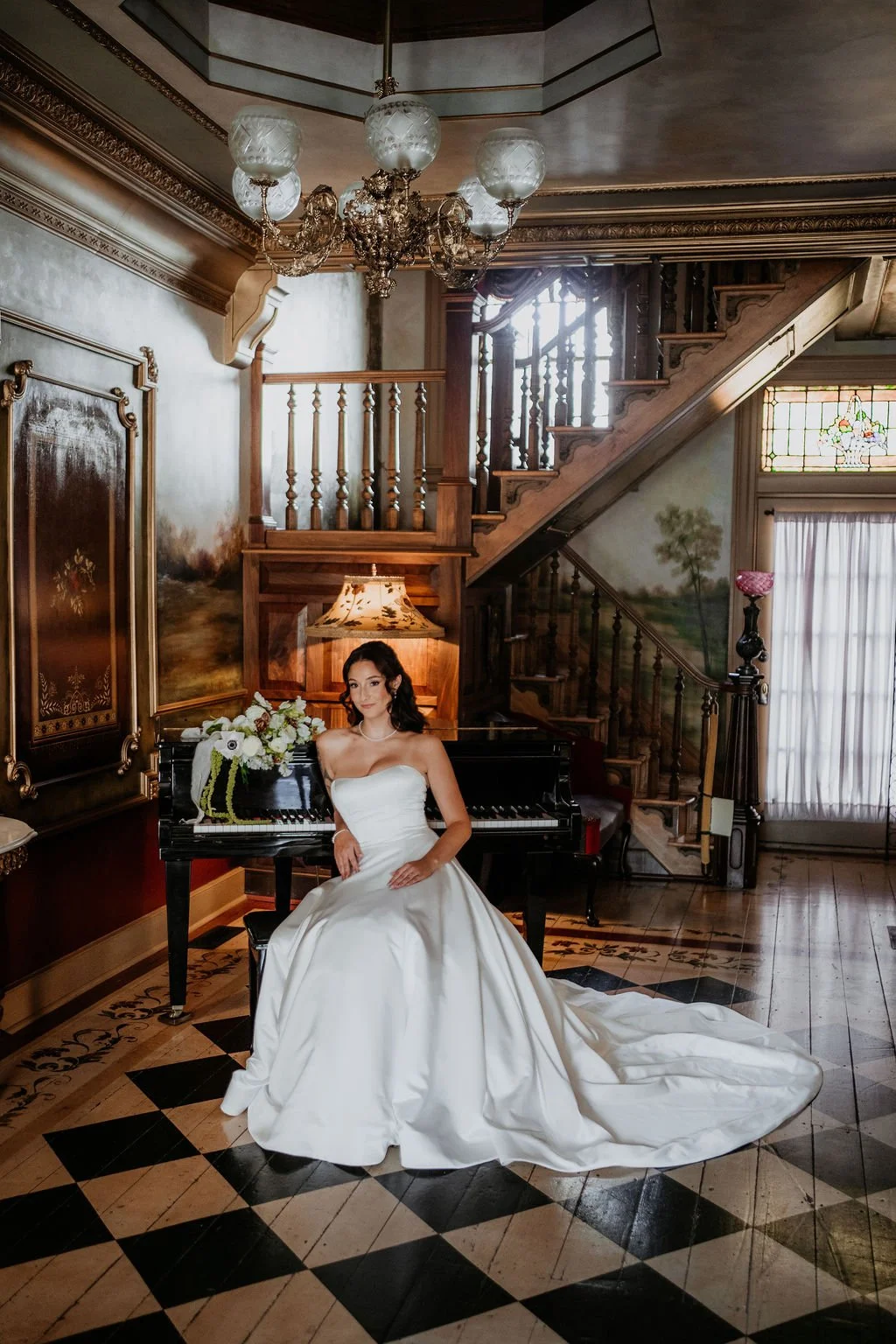 A woman in a white wedding dress sits beside a black grand piano decorated with a flower arrangement, in a richly decorated room with wood-paneled walls, a staircase, and stained glass windows.