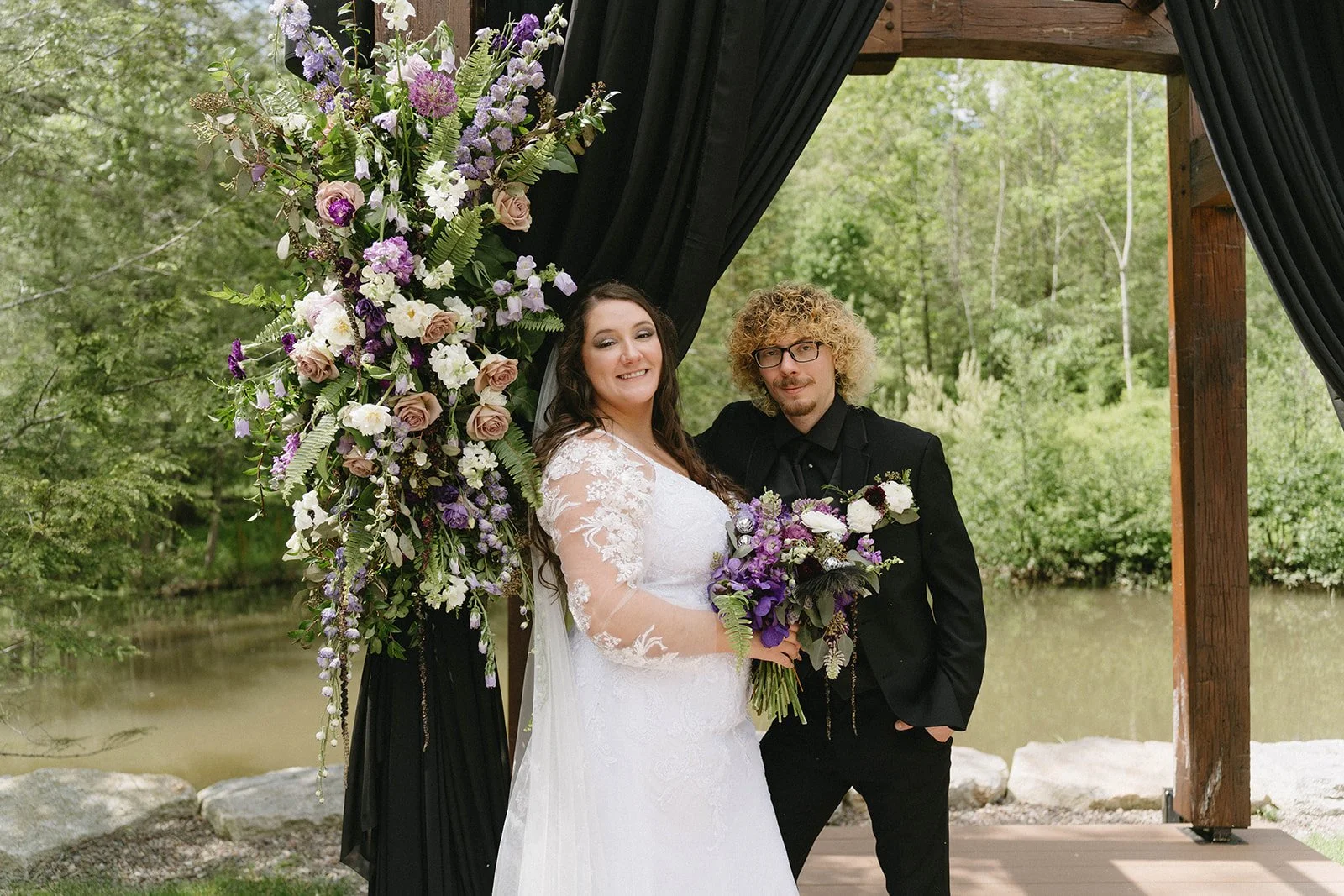 A bride and groom pose under a decorated outdoor arch with purple, white, and pink flowers, with a green forest and pond in the background.