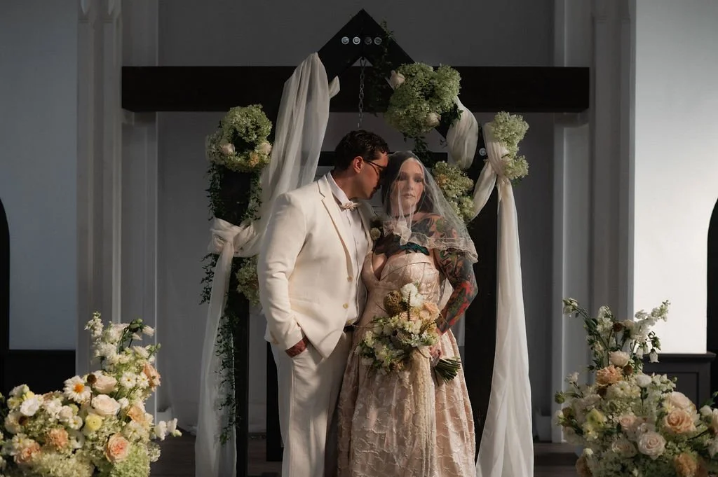 A couple dressed in wedding attire standing together at a wedding ceremony with floral decorations and an arch behind them.