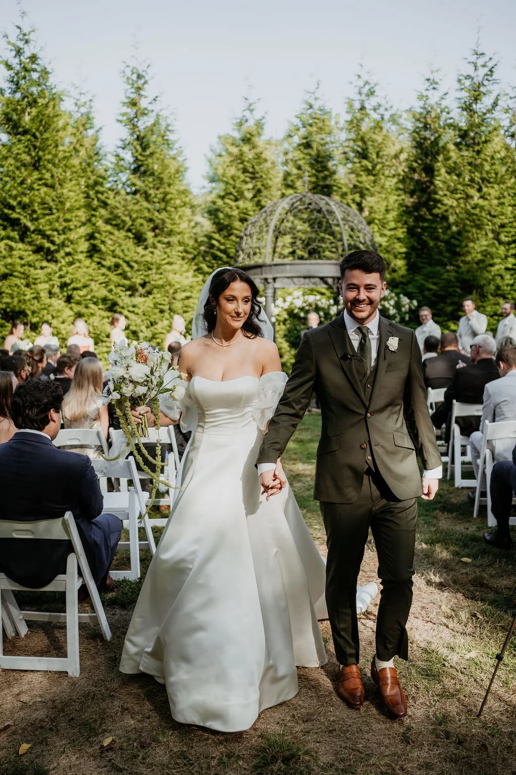 A bride and groom walking hand in hand at their outdoor wedding ceremony. The bride wears a white off-the-shoulder wedding gown, and the groom wears a dark suit and brown shoes. Guests are seated on white chairs in the background with a lush green fo