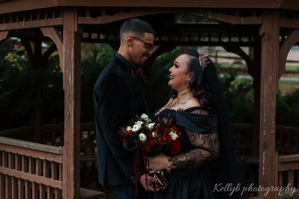 A bride and groom are standing close to each other under a wooden gazebo, smiling and looking into each other's eyes. The bride is wearing a black dress with lace sleeves, a veil, and holding a bouquet of red, black, and white flowers. The groom is i