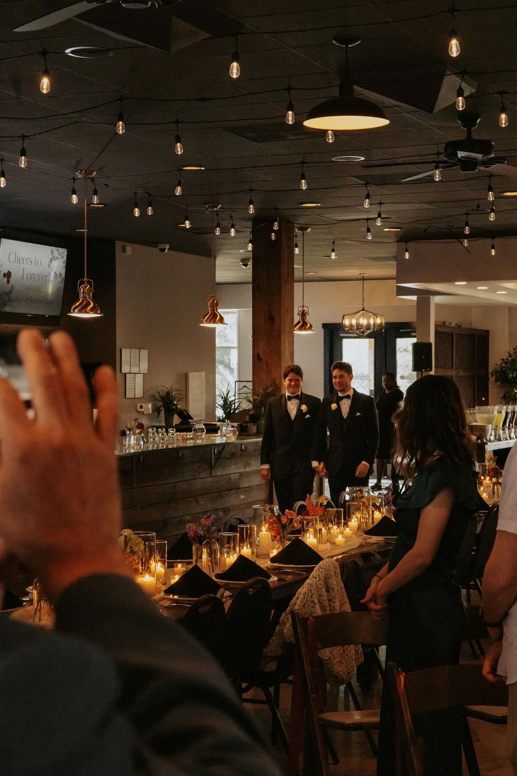 Two men in tuxedos holding hands at a wedding reception, with guests seated at decorated tables with candles and floral arrangements in a warmly lit venue.