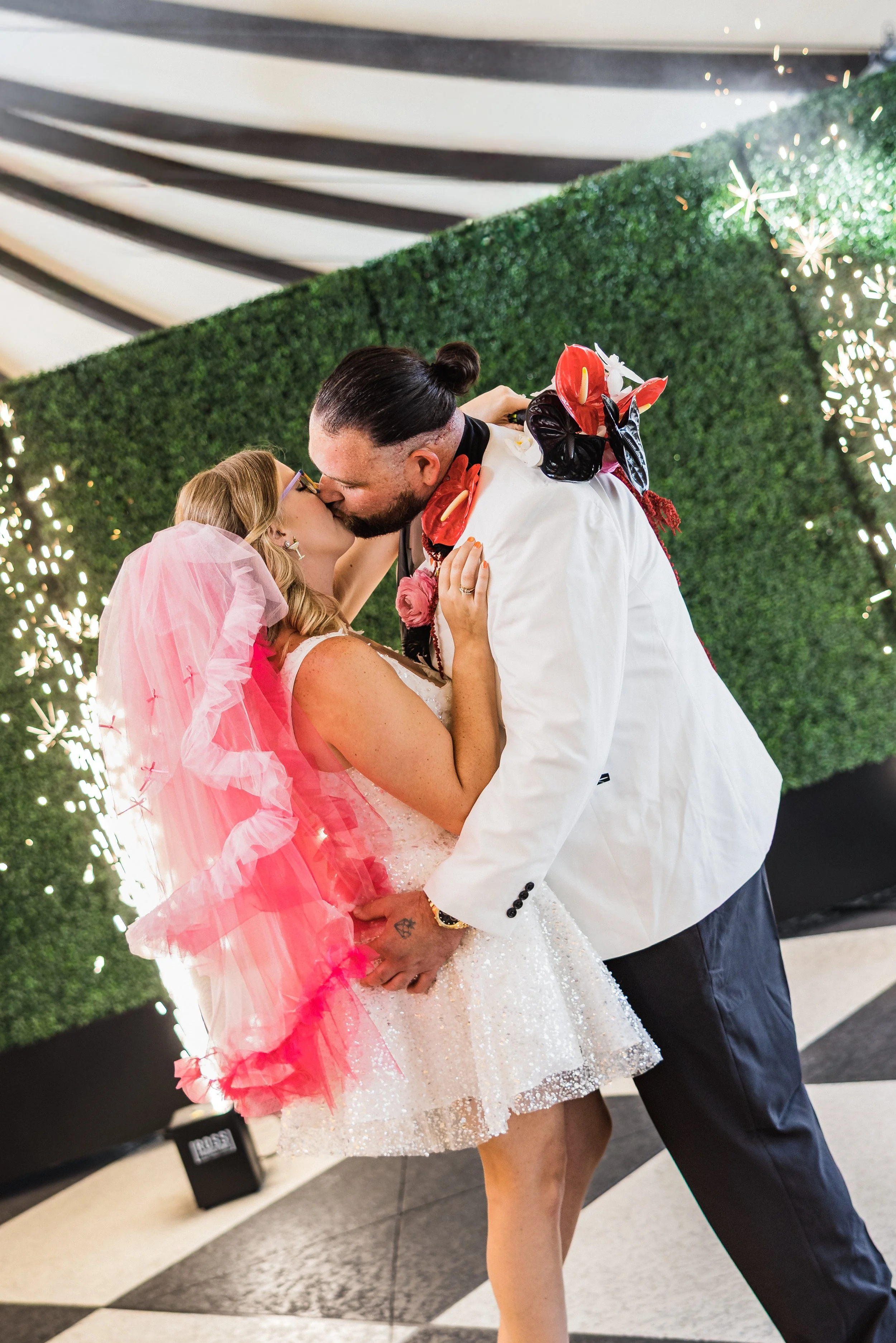 A newlywed couple shares a kiss at their wedding reception, with the bride wearing a sparkly white dress and a pink veil, and the groom in a white suit with floral decorations, against a green hedge backdrop with sparklers.