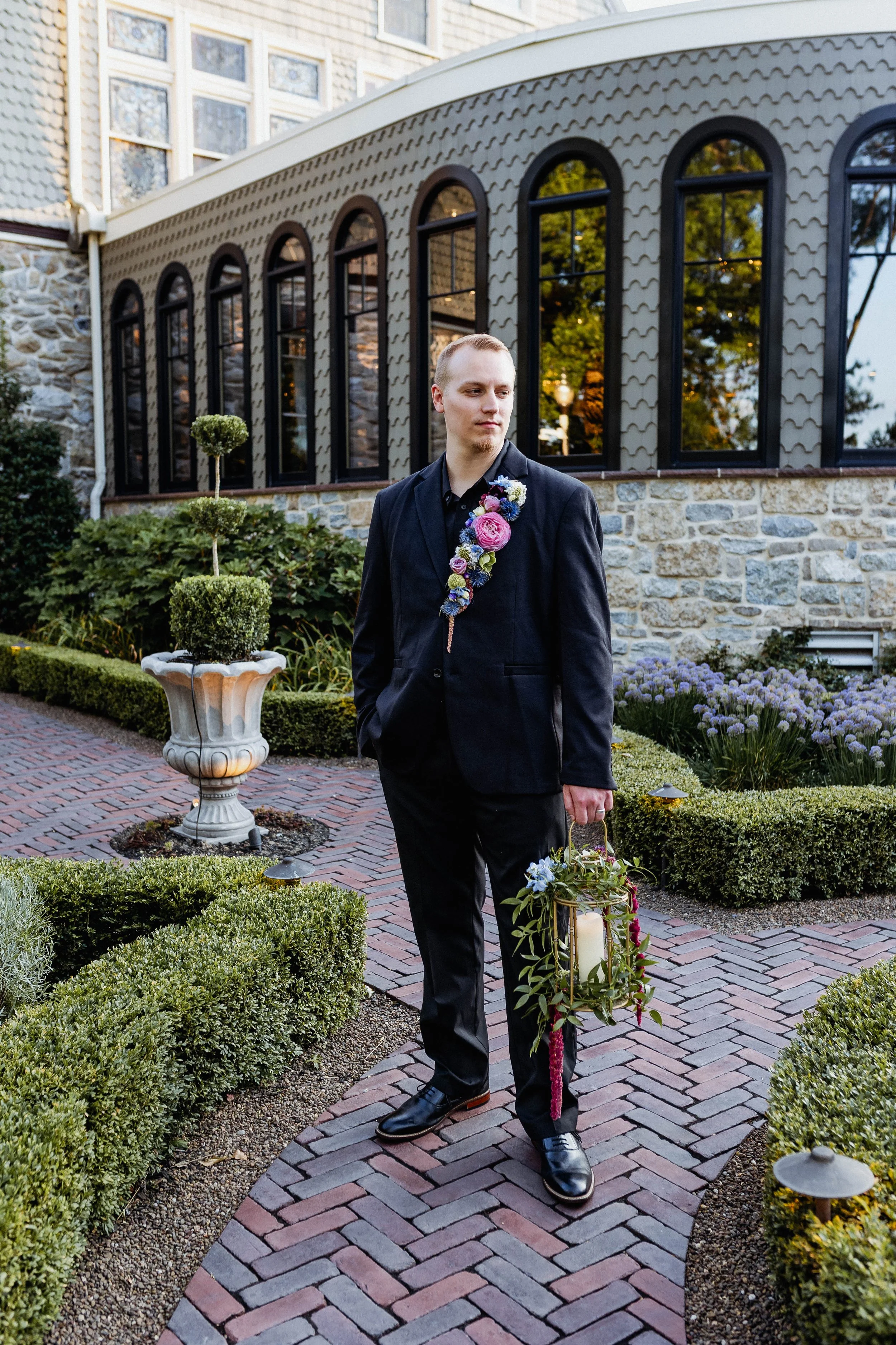 A young man in a black suit with a floral sash and holding a lantern with a candle and flowers, standing on a brick pathway in a garden with neatly trimmed bushes, purple flowers, and a stone building with large arched windows in the background.