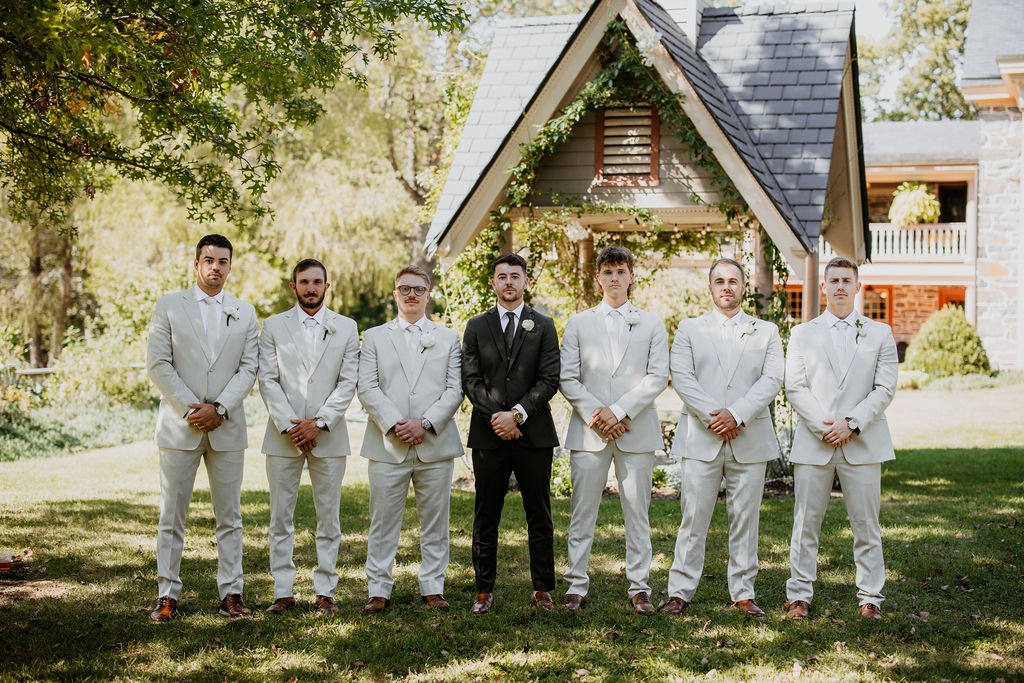 Group of seven men dressed in suits standing outdoors in front of a small house with trees and a building in the background.