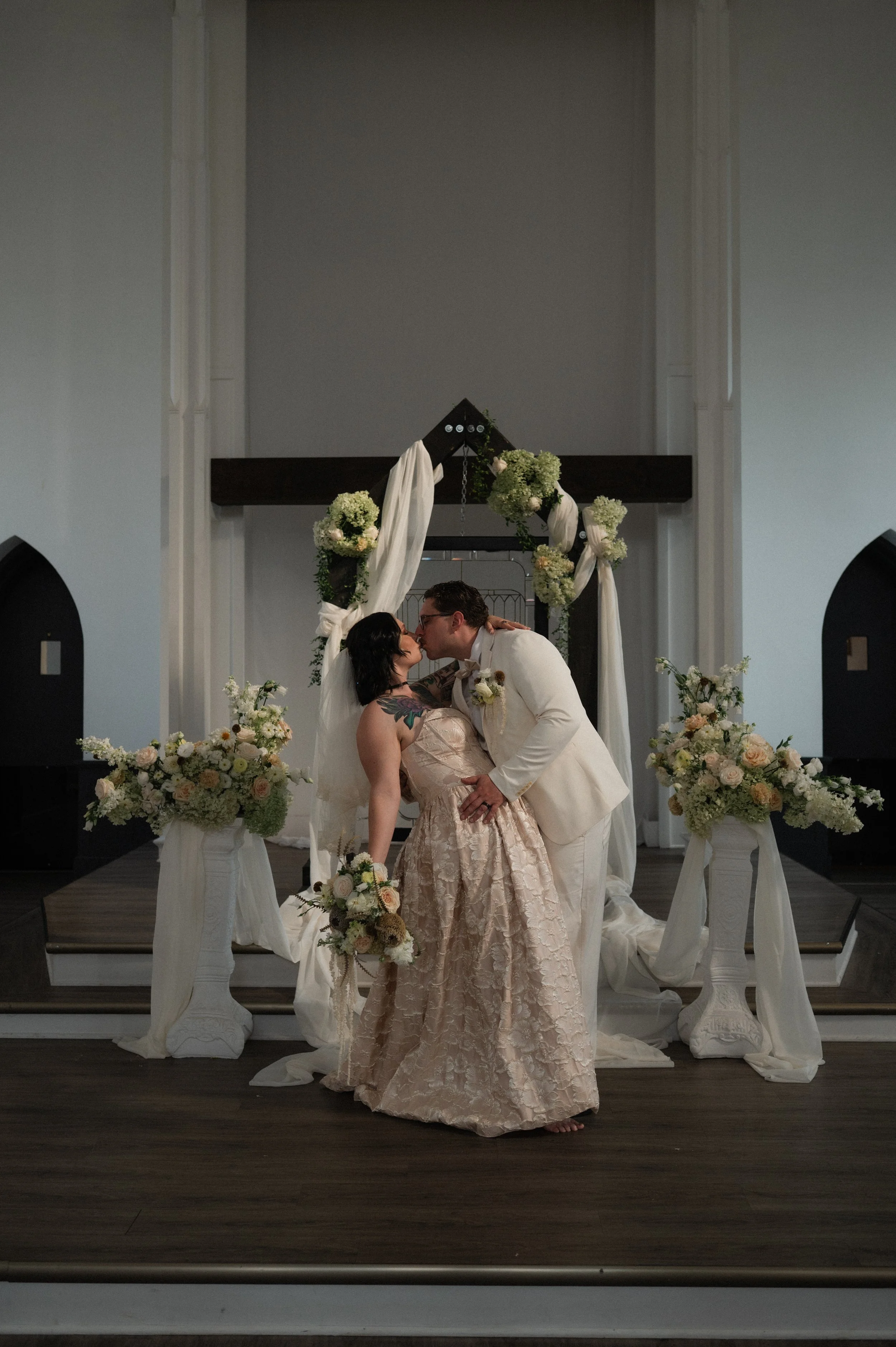 A couple in wedding attire kissing in front of their wedding altar decorated with flowers and drapes.