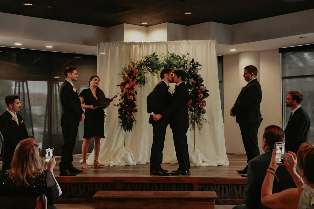 A same-sex couple kissing during a wedding ceremony on a small stage, with an officiant and four witnesses, in a room with large windows. Guests are capturing the moment on their phones.