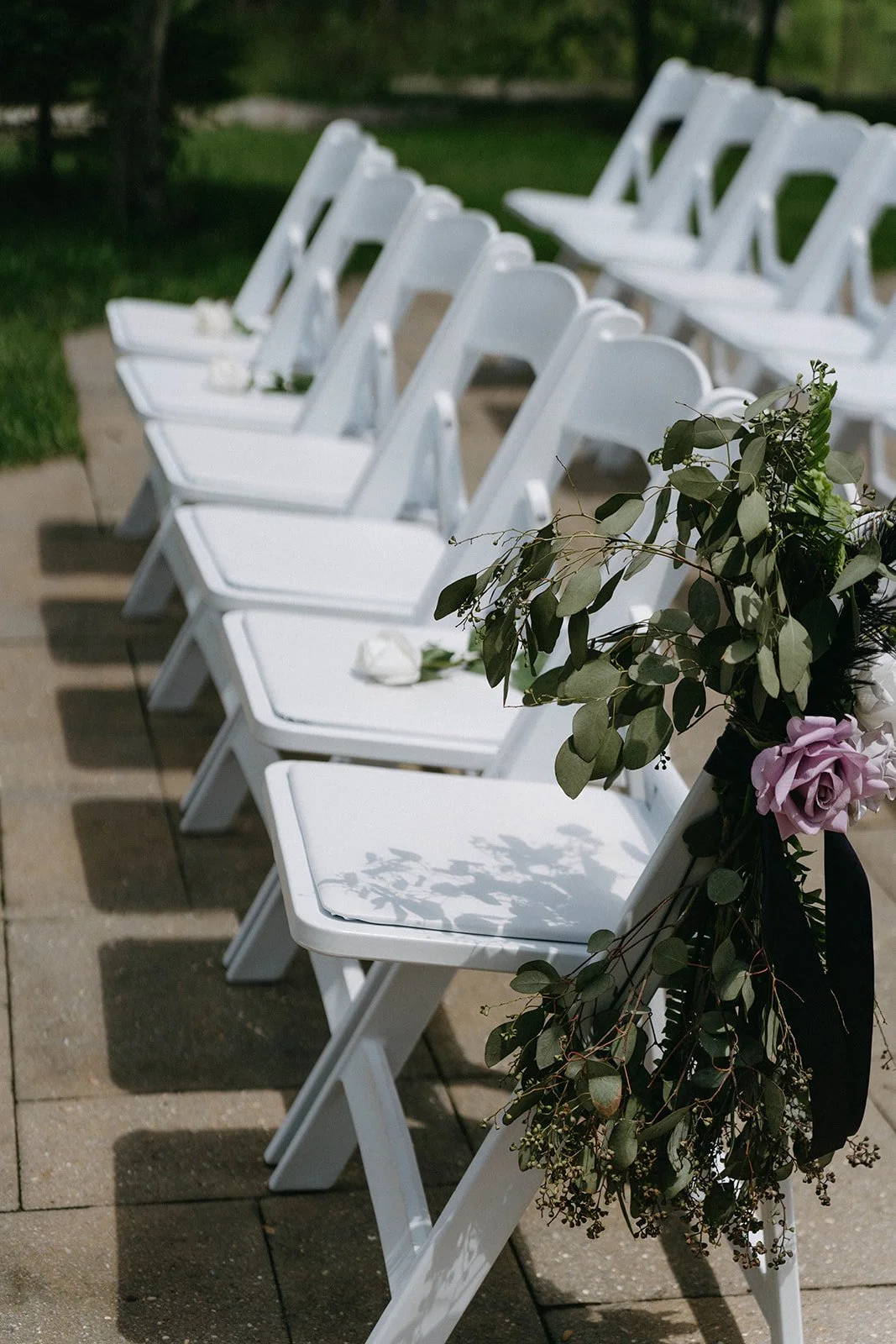 White folding chairs decorated with greenery and pink flowers, arranged outdoors for a wedding ceremony.