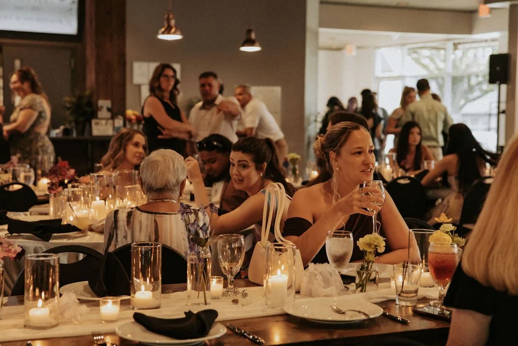 People socializing at a dinner party with decorated tables, candles, and drinks in a warmly lit venue.
