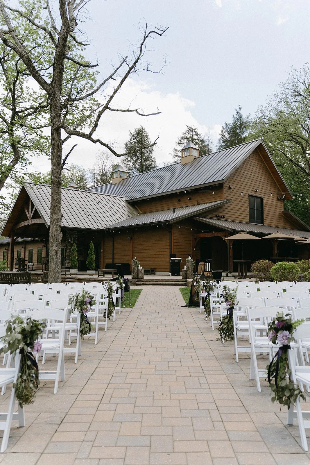 Outdoor wedding ceremony setup at a rustic barn with white chairs decorated with floral arrangements, a brick aisle, and umbrellas on the patio.