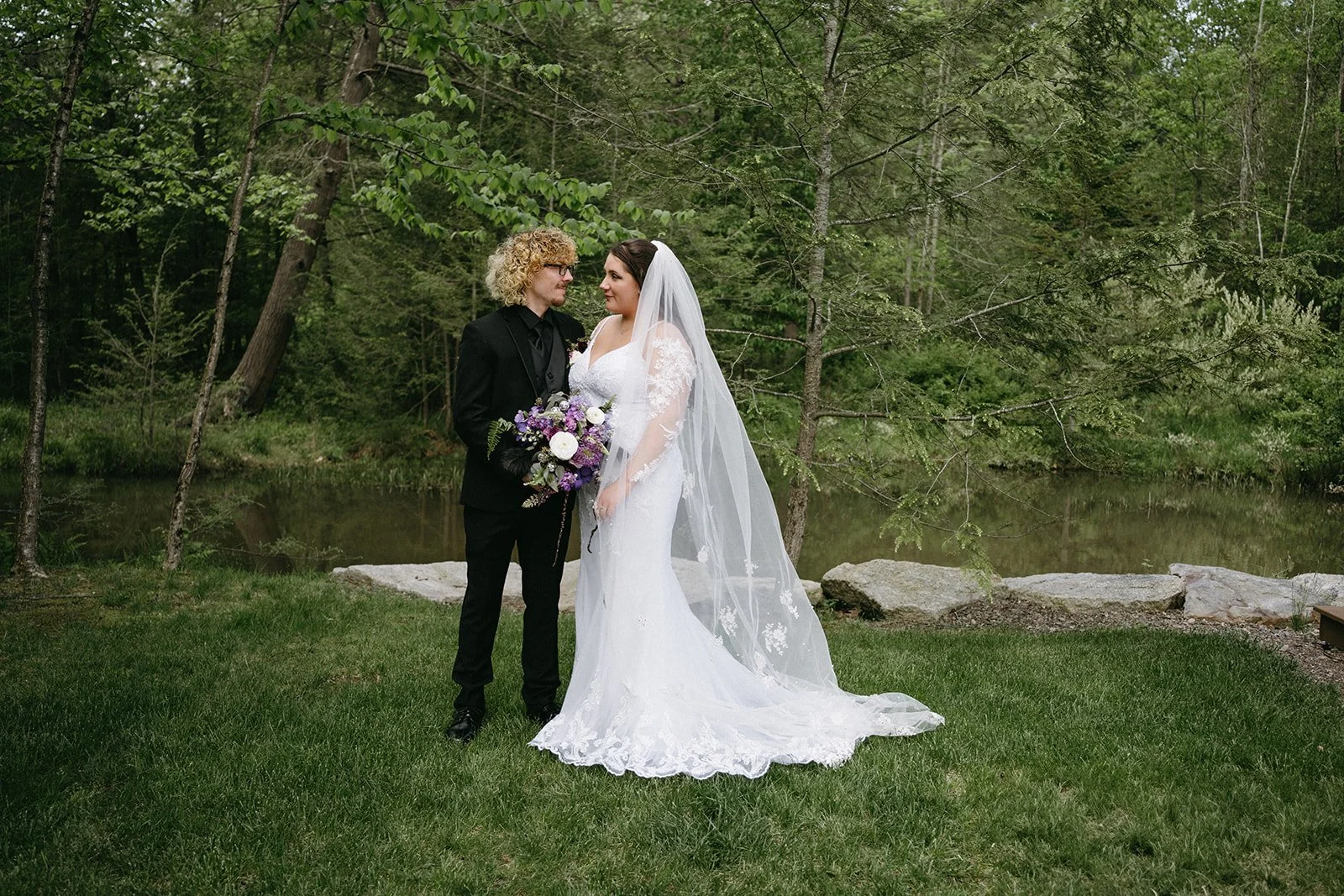 A bride in a white lace wedding dress with a veil holding a bouquet of purple, white, and green flowers, and a groom in a black suit standing close together by a pond and trees in a lush outdoor setting.