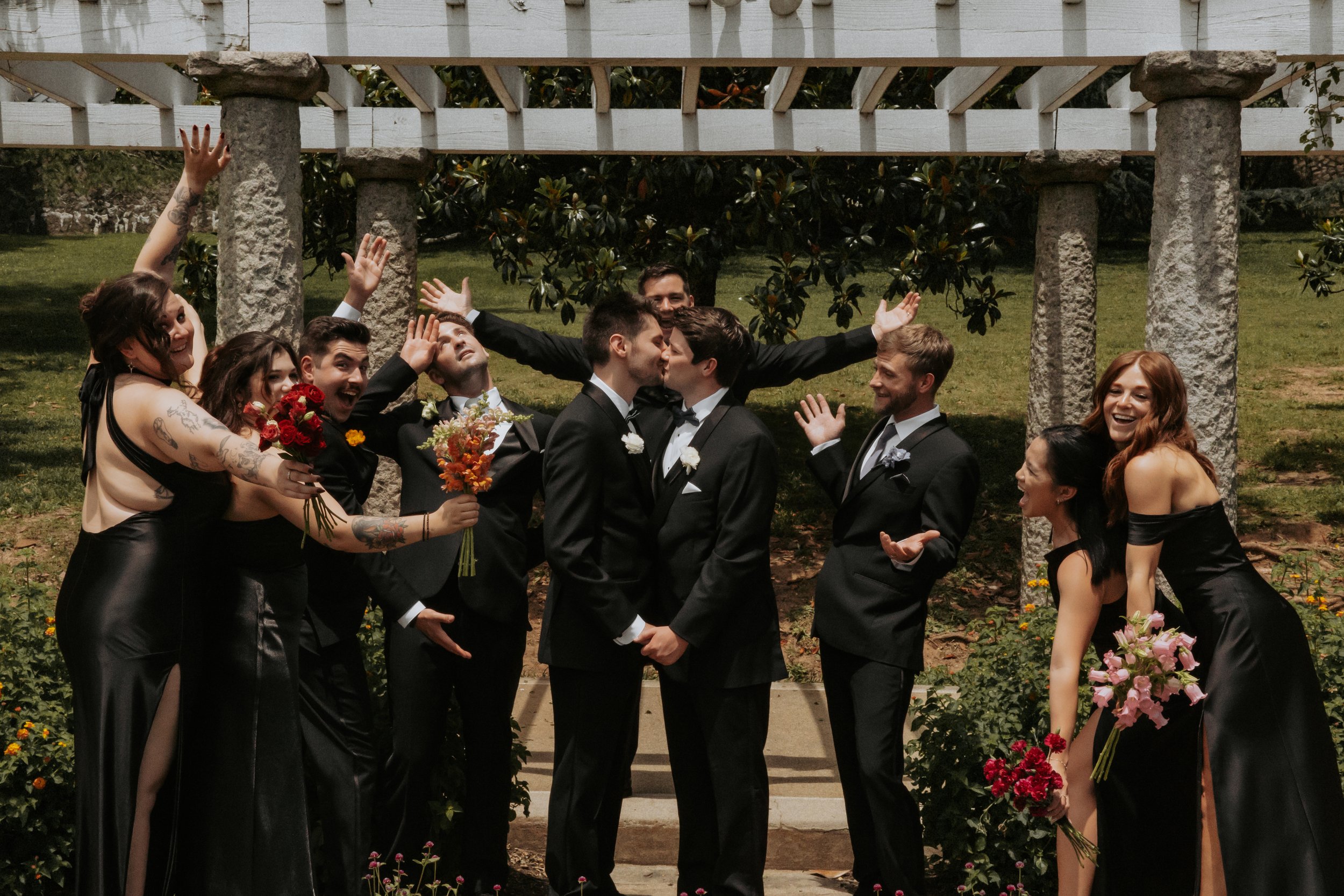 A wedding party with two grooms and their friends in formal attire, celebrating outdoors with flowers and greenery.
